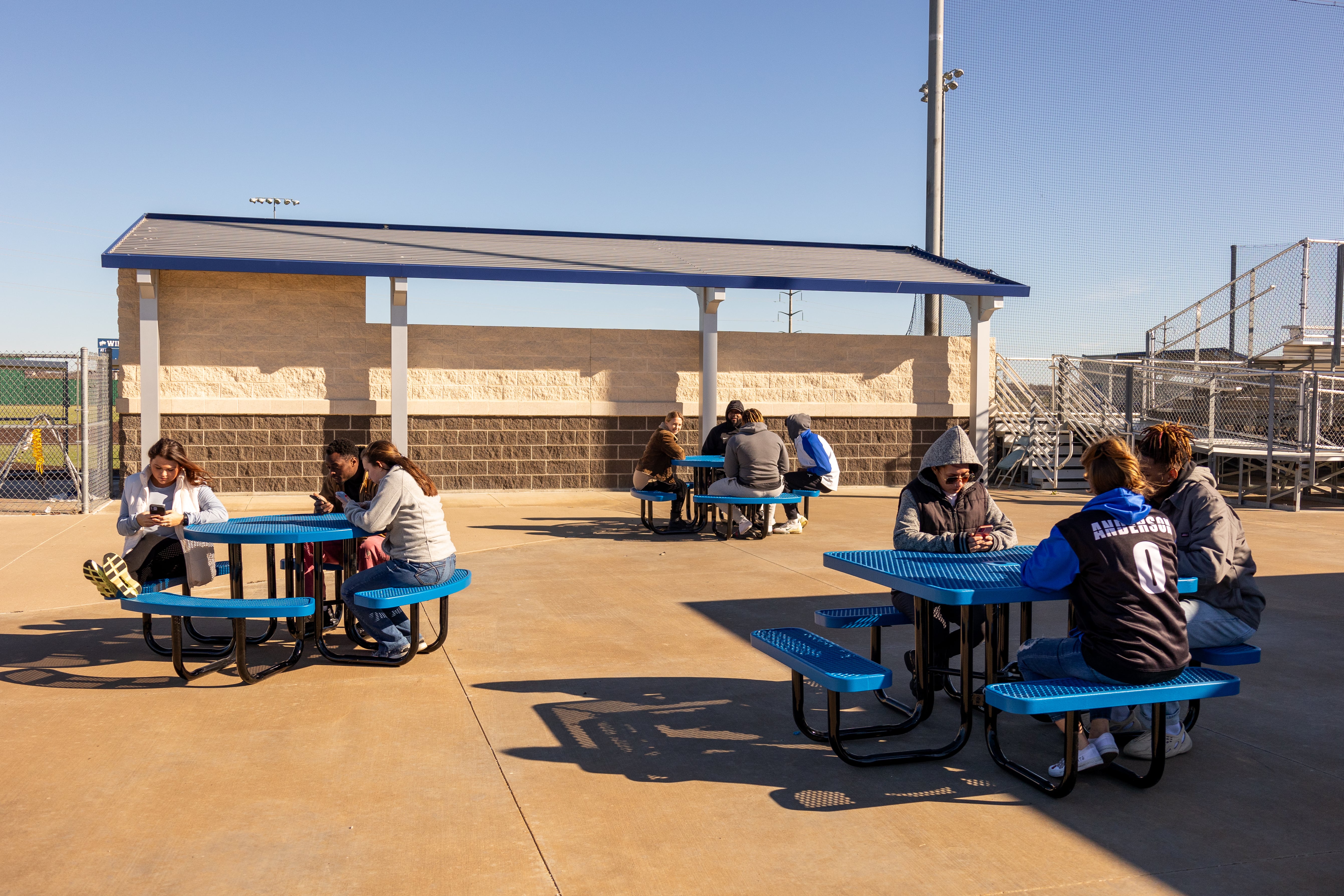 Multiple Blue MyTCoat Square Portable Picnic Tables with People Sitting in School with Football Field Bleachers Background