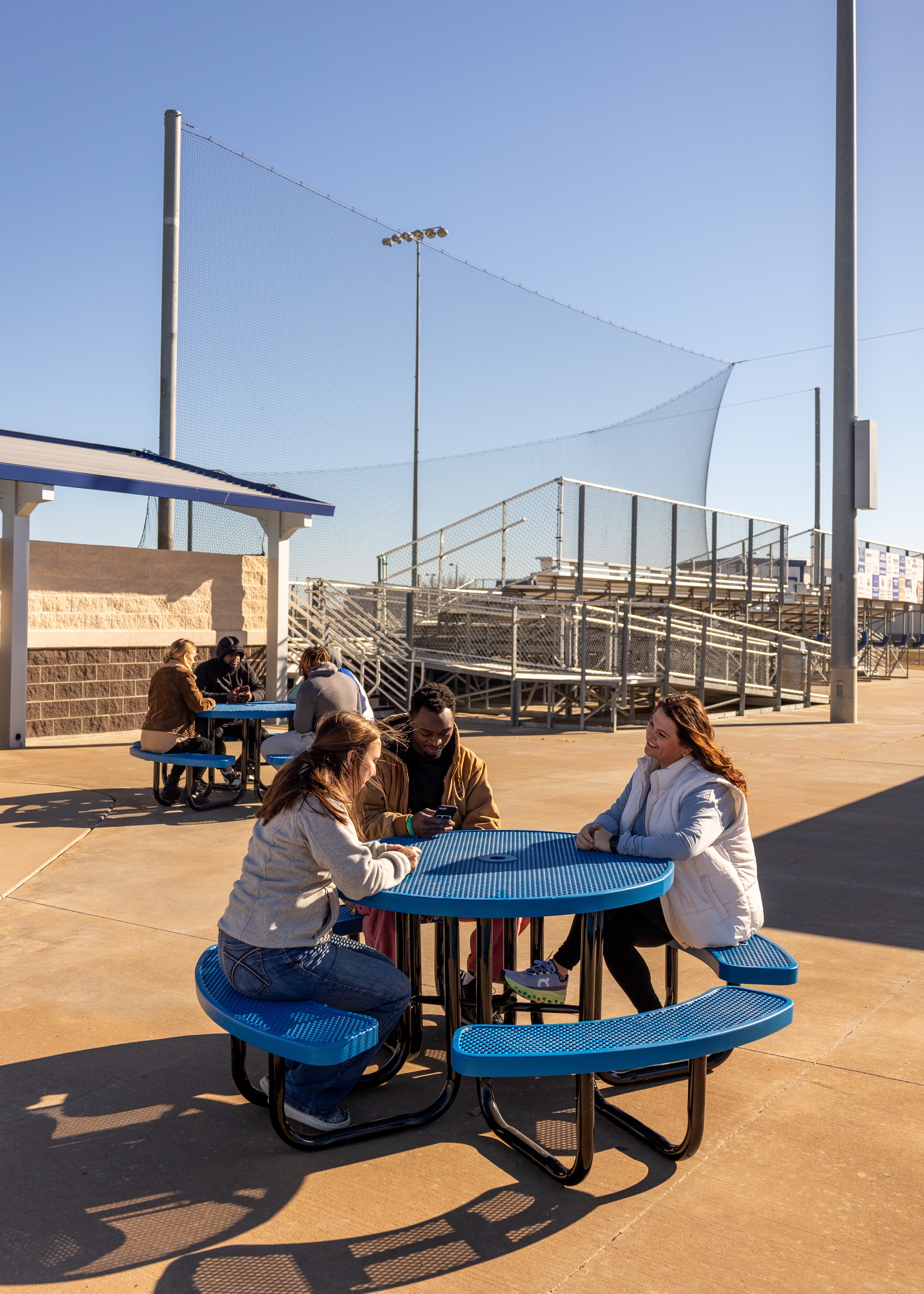 Two Blue MyTCoat Round Portable Picnic Tables Lifestyle Image in School Courtyard with People Sitting by Football Field