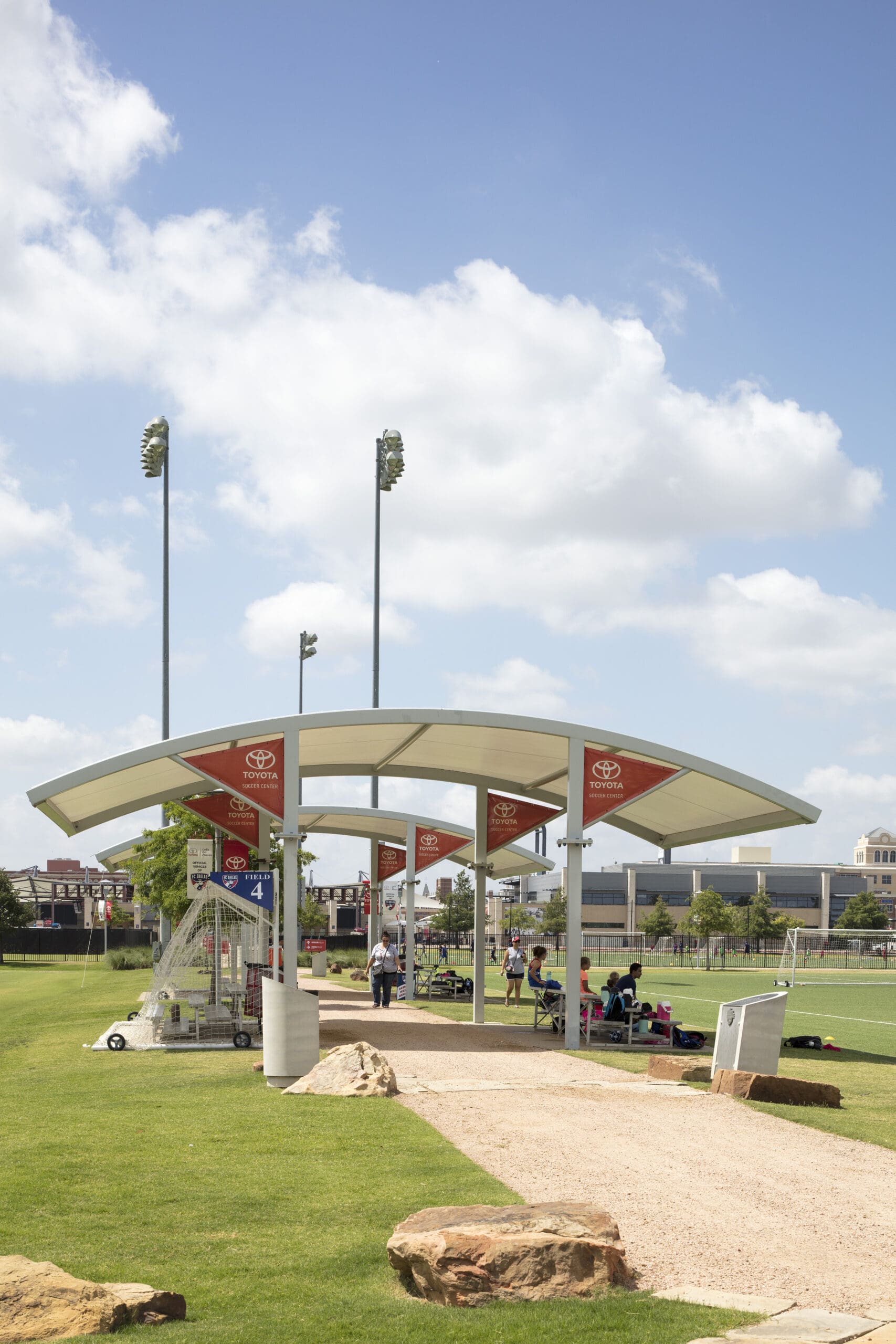 USA Shade Panorama Cantilever Covered area with sports equipment on a grassy field under a blue sky with clouds.