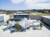 Outdoor area with white umbrellas and USA Shade Orion Shades and tables in front of a modern building with a clear sky.