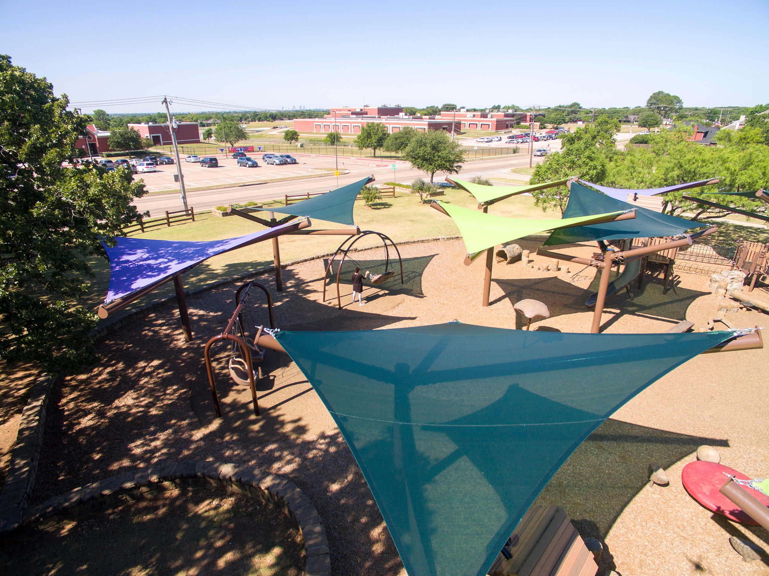 Playground with colorful USA Shade Arbor Shade sails and a view of a parking lot and buildings in the distance.