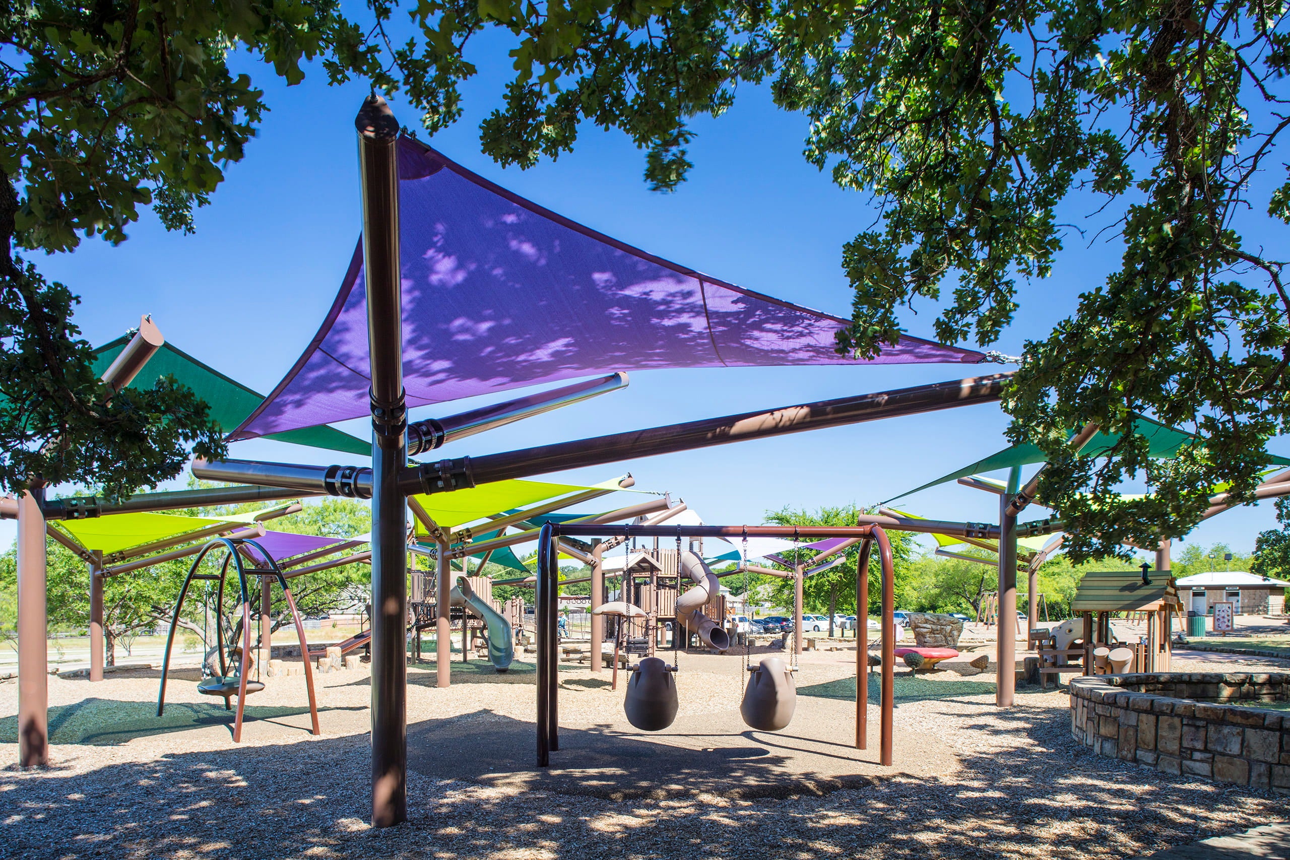 Children's playground with purple USA Shade Arbor Shade sails and wooden play structures under a blue sky.