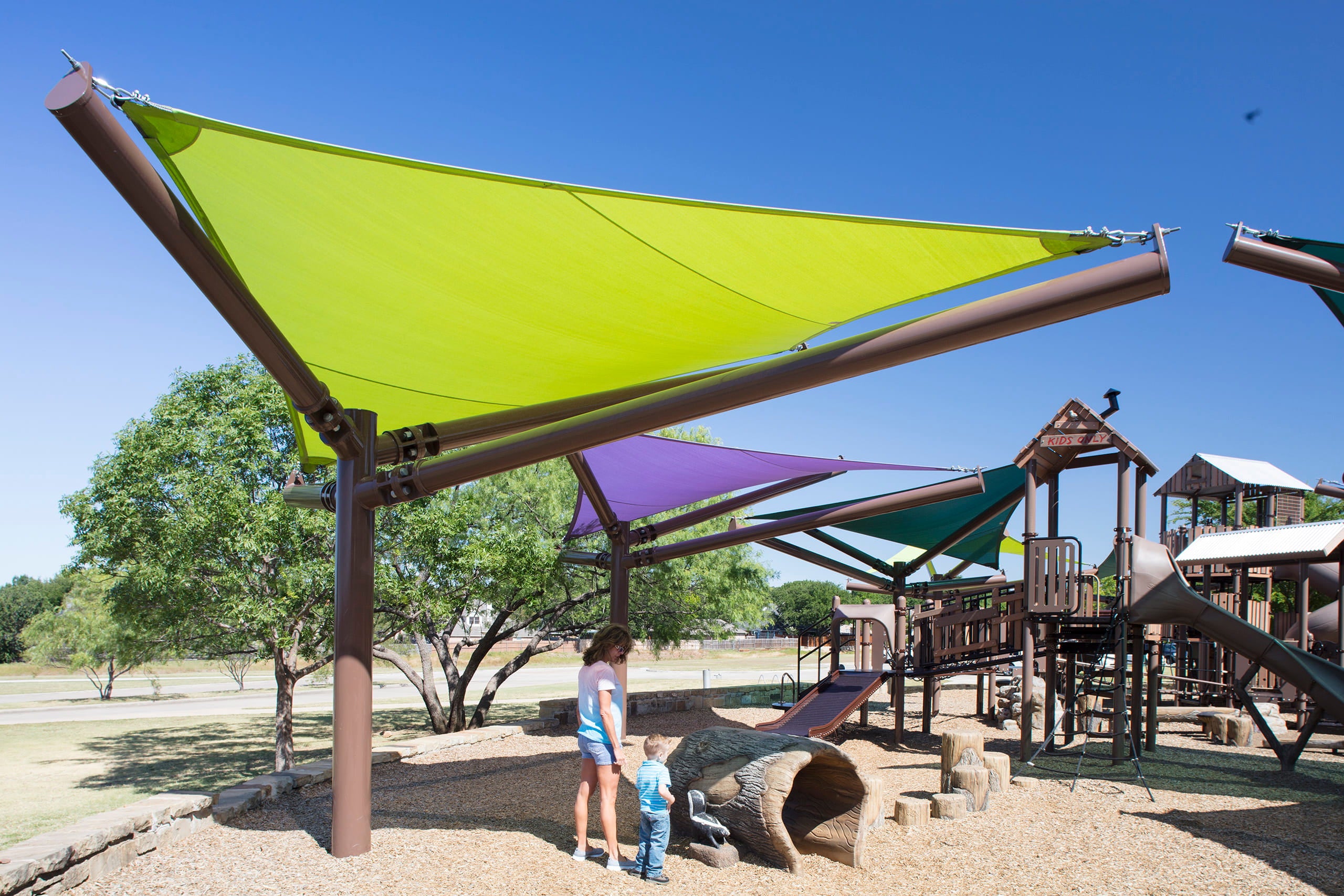 Children playing under a green USA Shade Arbor Shade sail at a playground with a clear blue sky.