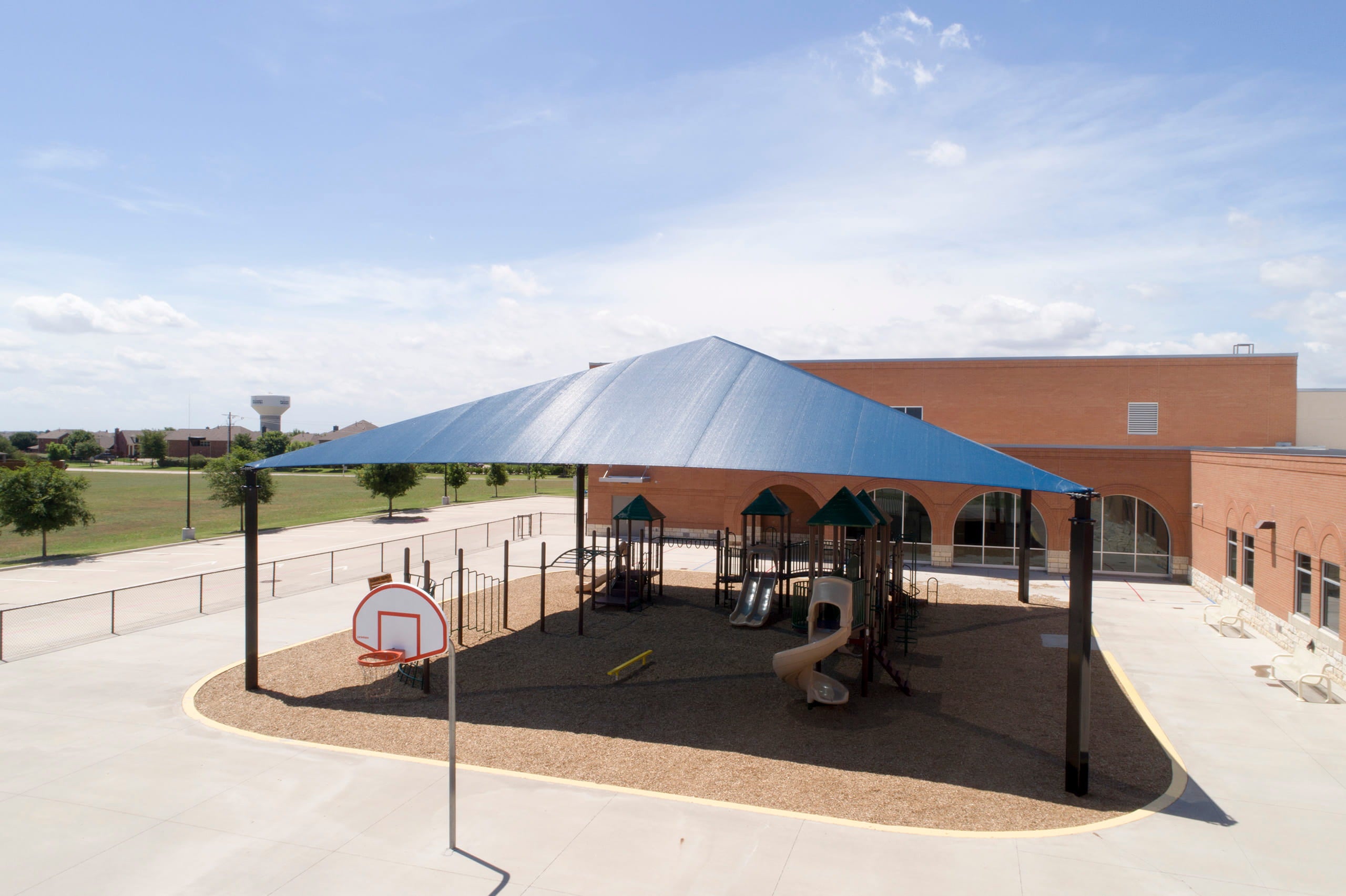 Playground with blue USA Shade Super Span Hip and basketball hoop in front of a building
