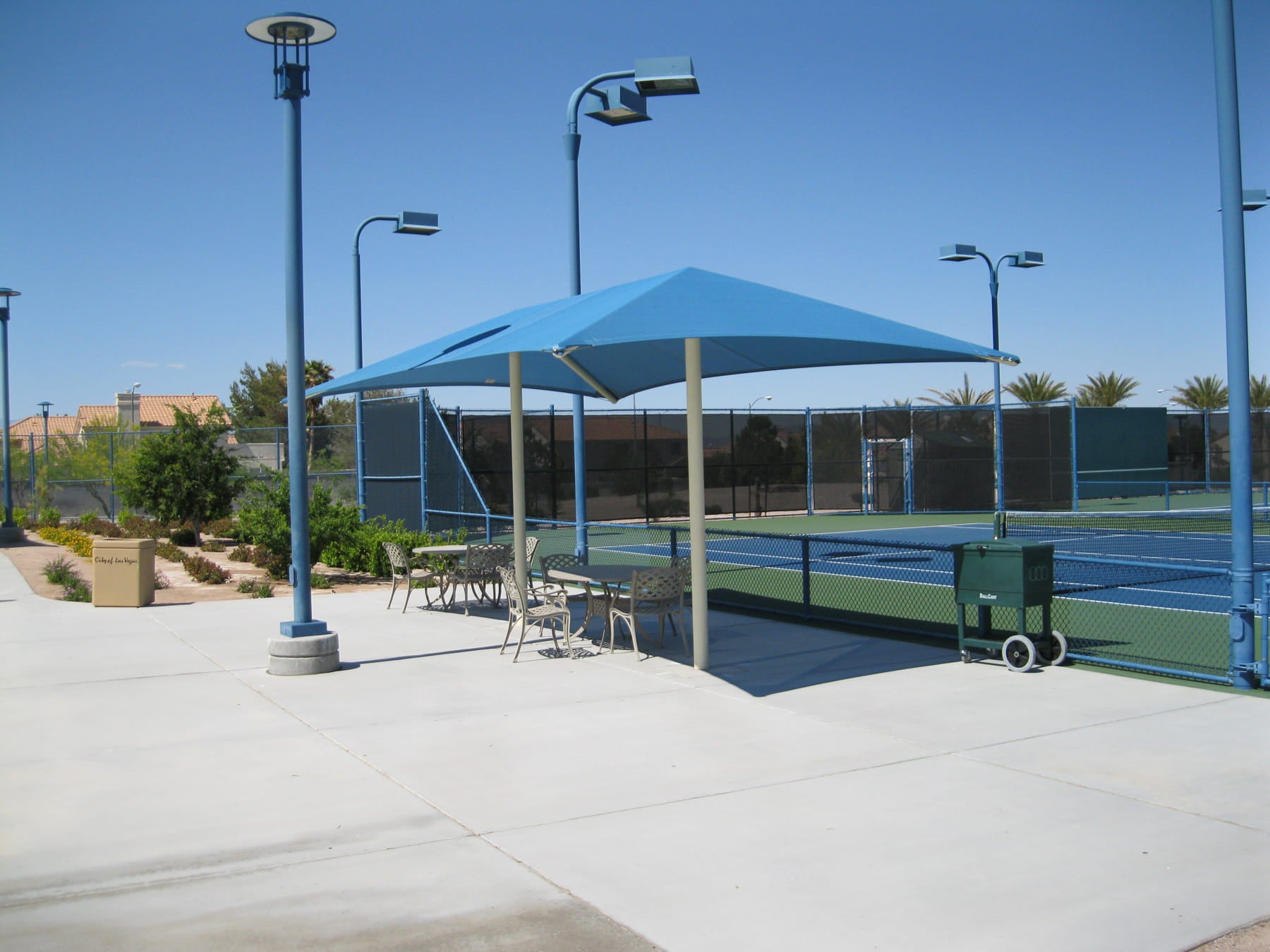 Tennis court with a blue USA Shade Two Post Hip Shade Structure and surrounding area on a clear day.