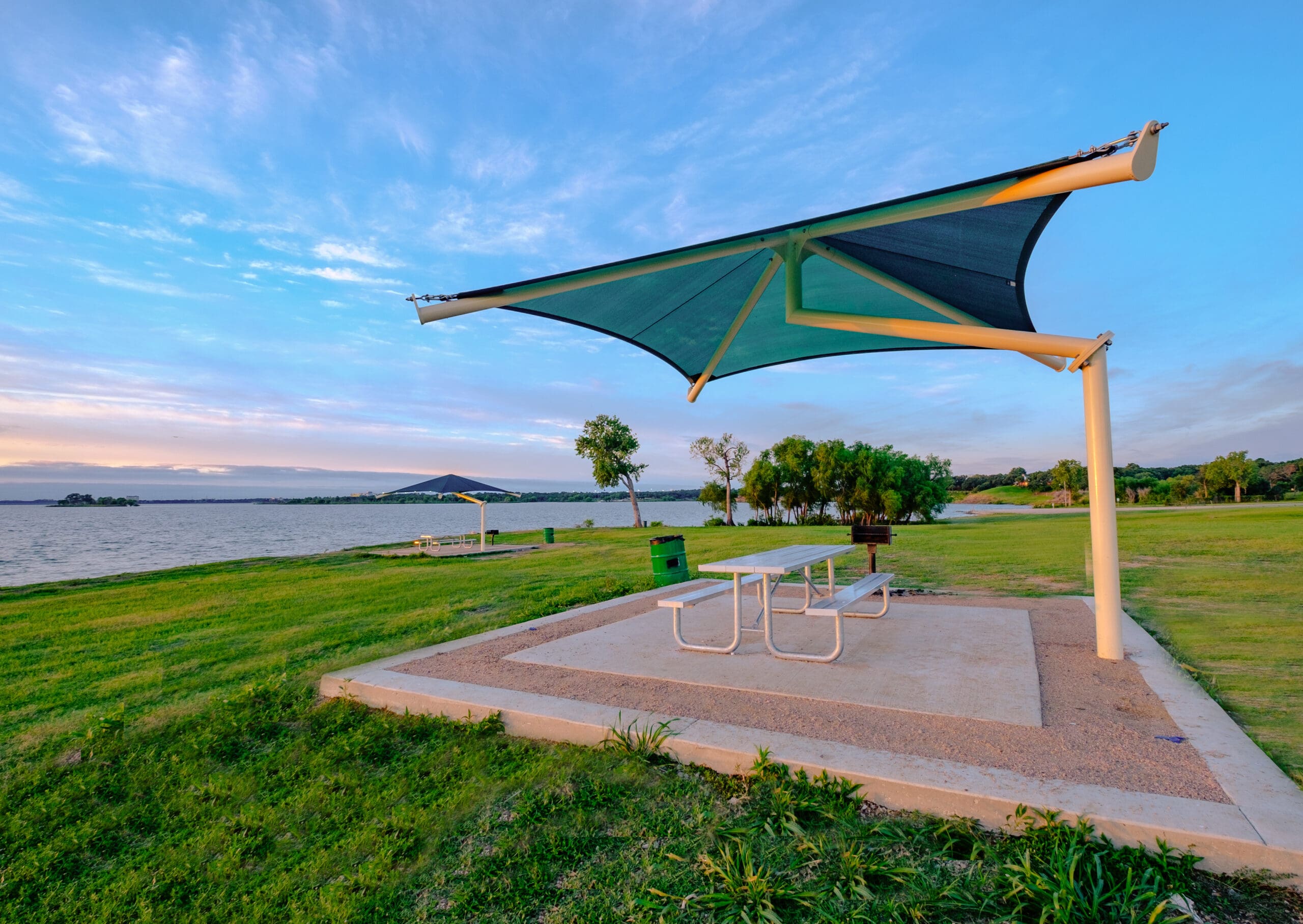Outdoor setting with a large green and beige USA Shade Single Post Cantilever Shade Structure over a picnic table by a lake.