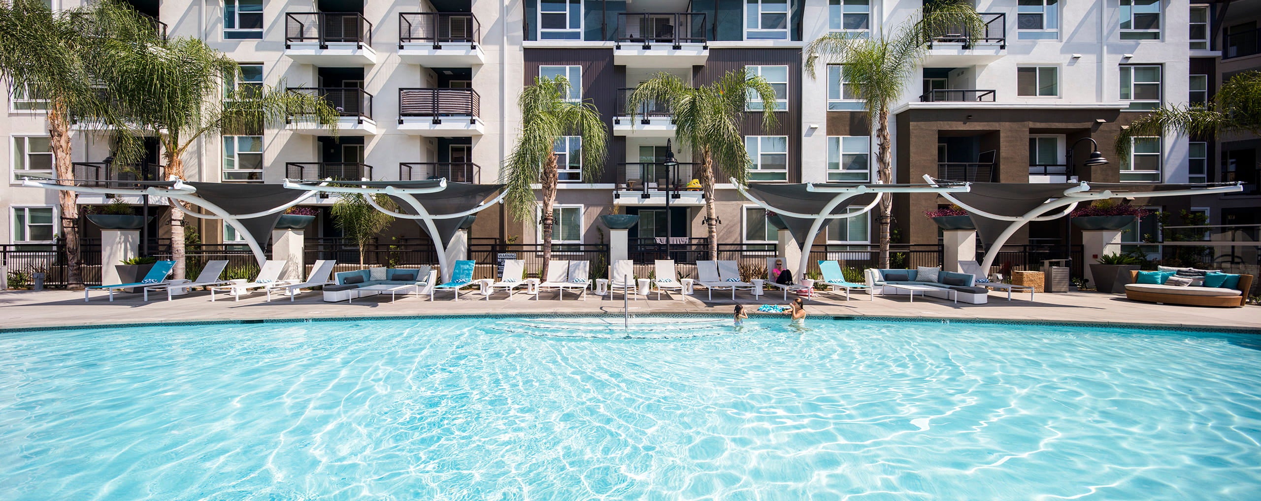 Pool area with lounge chairs and USA Shade Pegasus Shades in front of a multi-story apartment building.