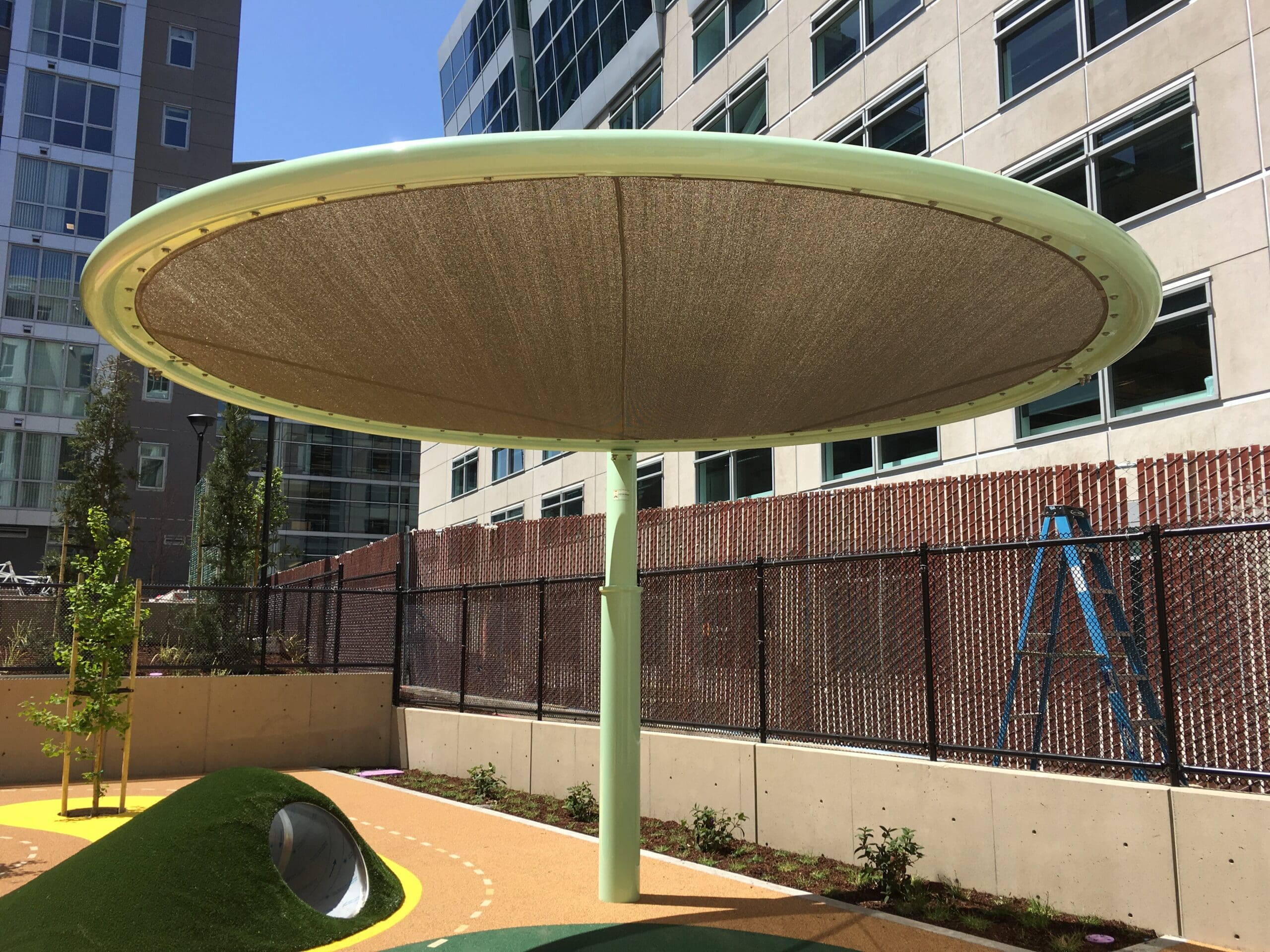 Round USA Shade Cantilever Disc Shade structure with a green frame and beige Fabric in an urban setting.