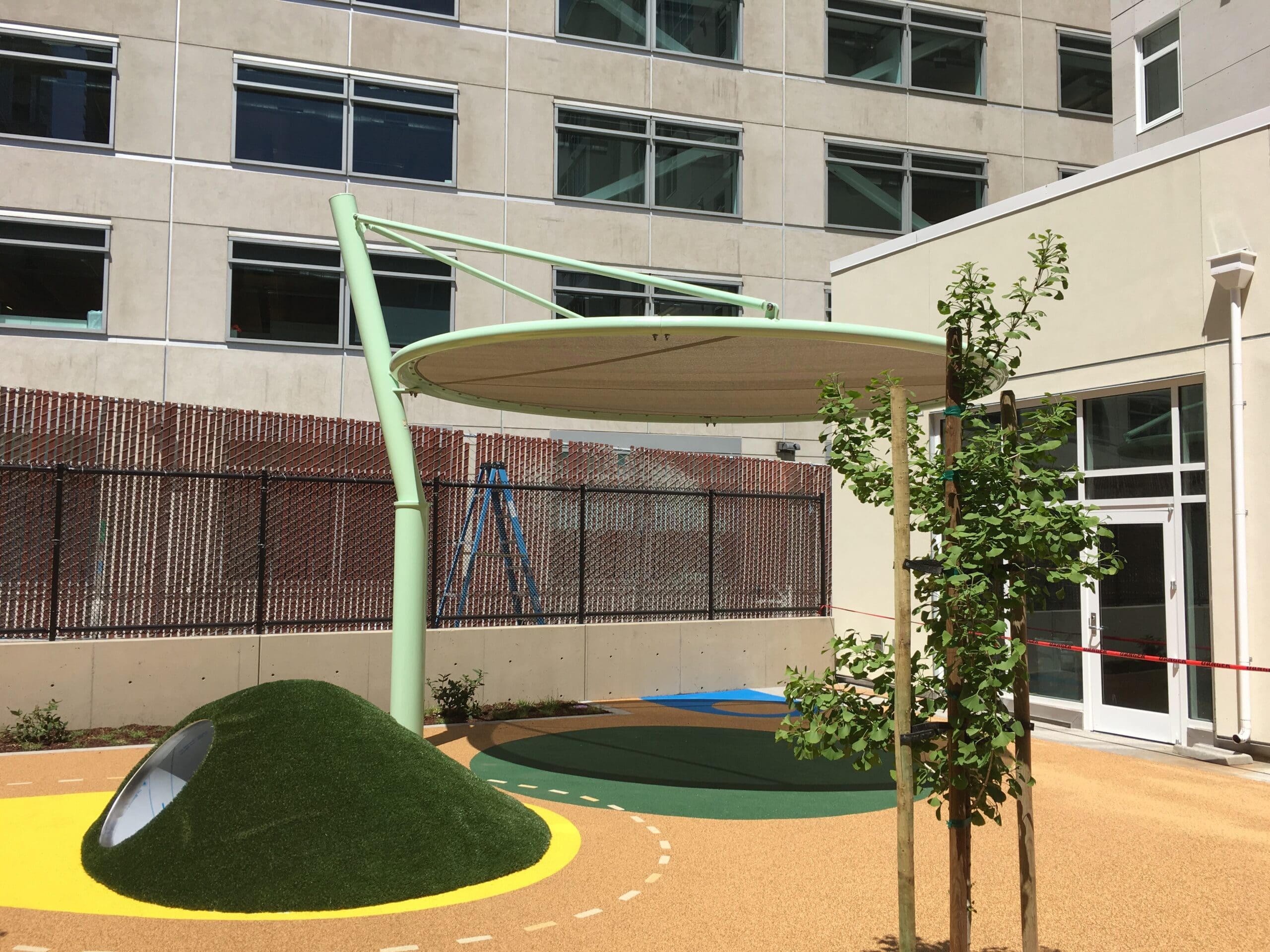 Children's playground area with green USA Shade Cantilever Disc shade and a building in the background