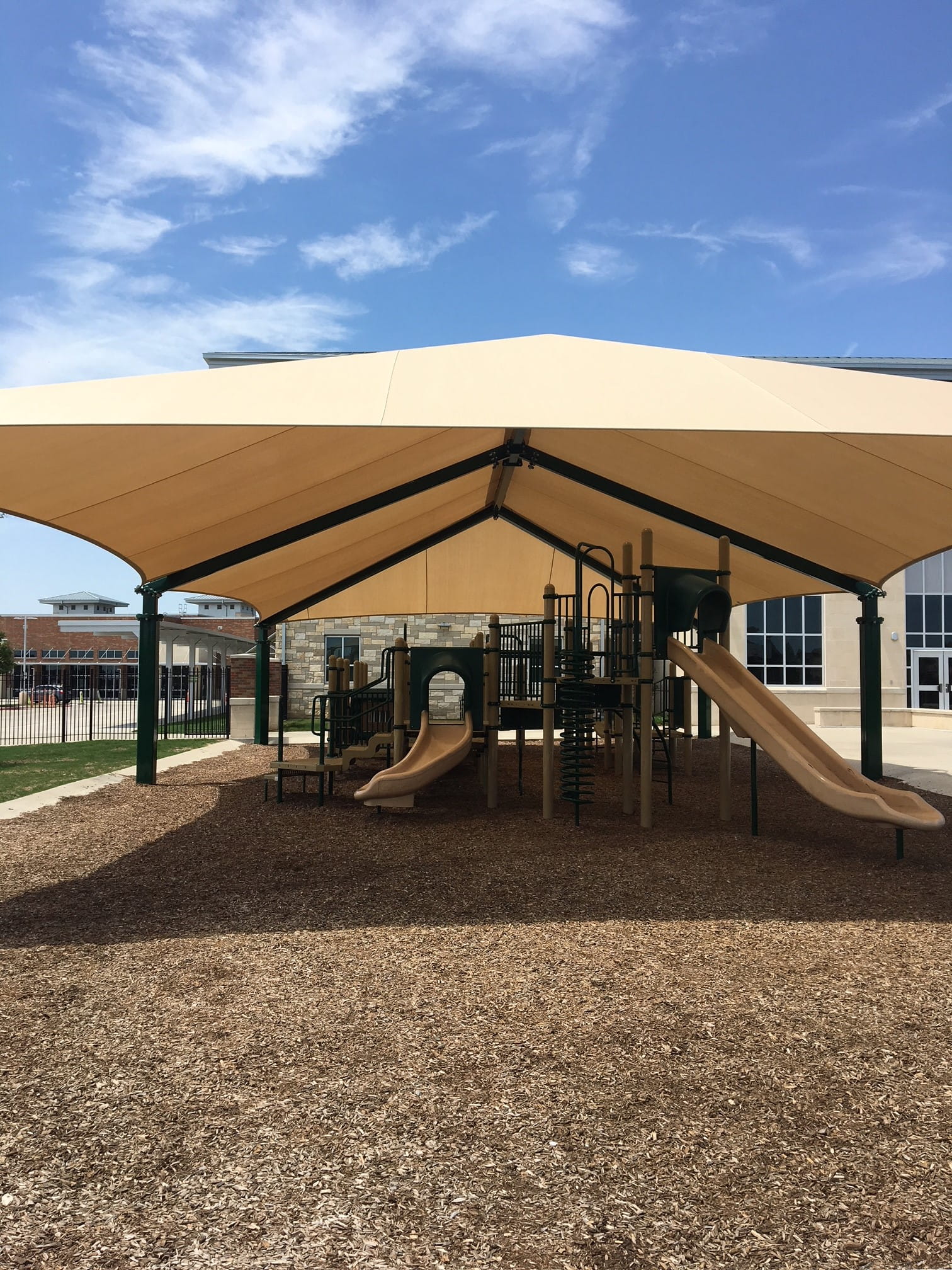 Playground with slides and climbing structures under a large beige USA Shade Super Span 6-Post Hip on a sunny day.
