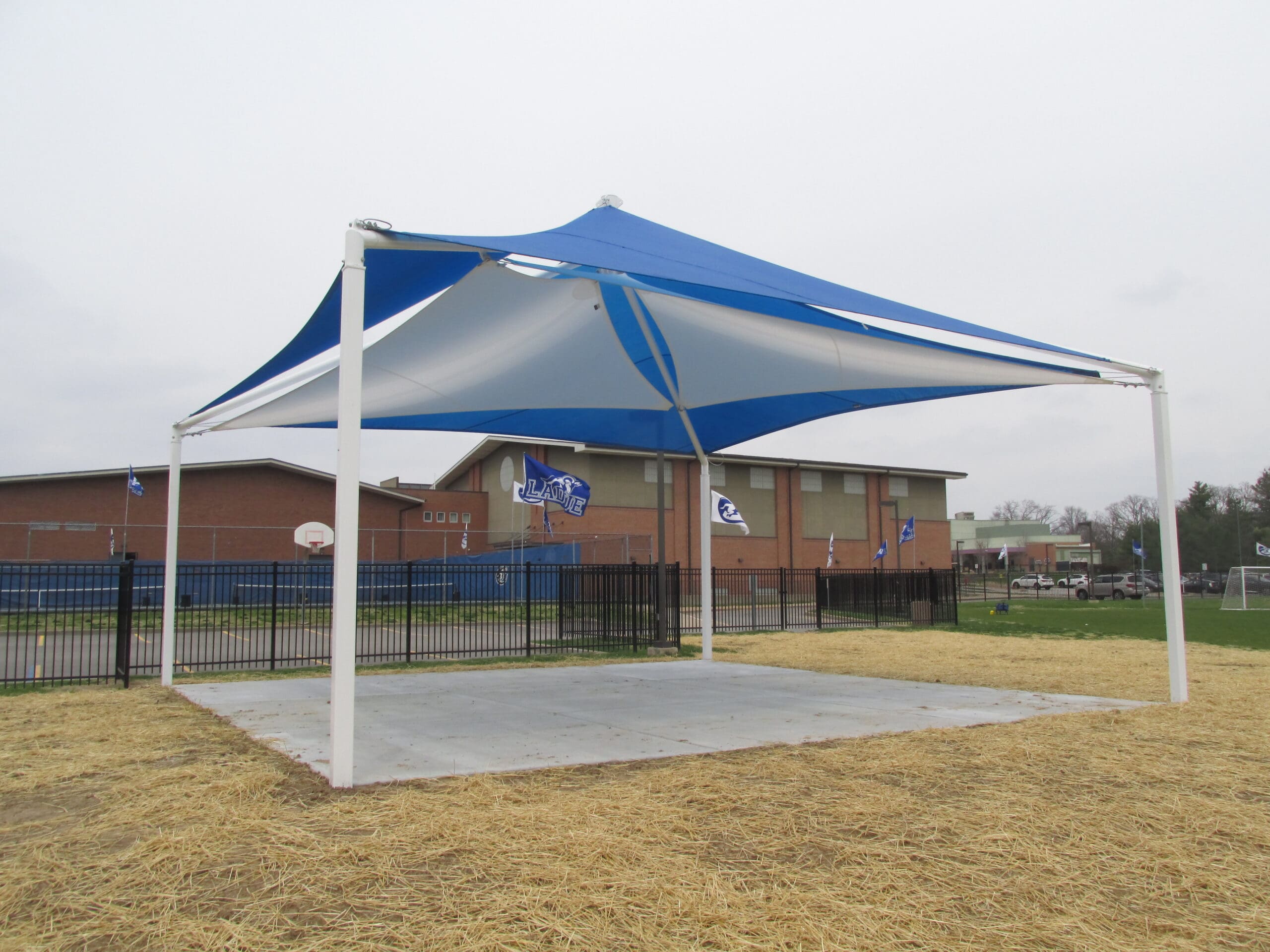 Blue and white USA Shade Mariner Pyramid Shade structure on a grassy area with a building in the background