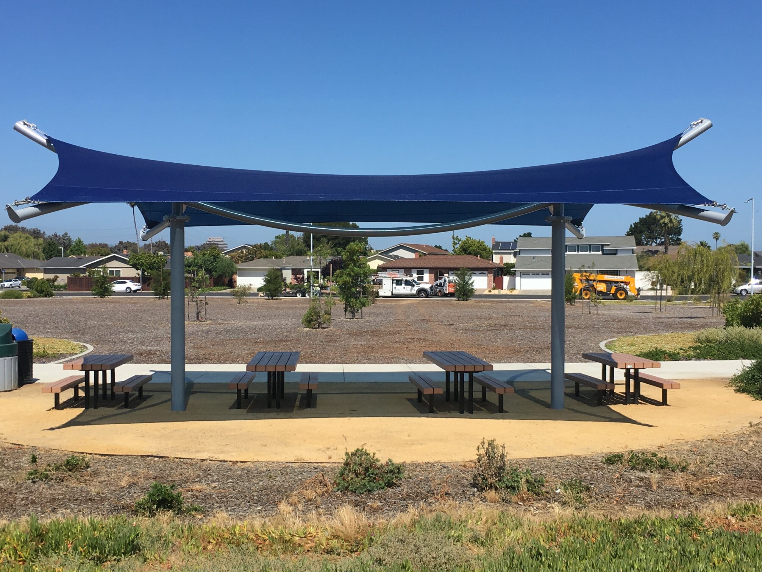 Blue USA Shade Aquarius Shade structure over picnic tables in a park setting with houses in the background.