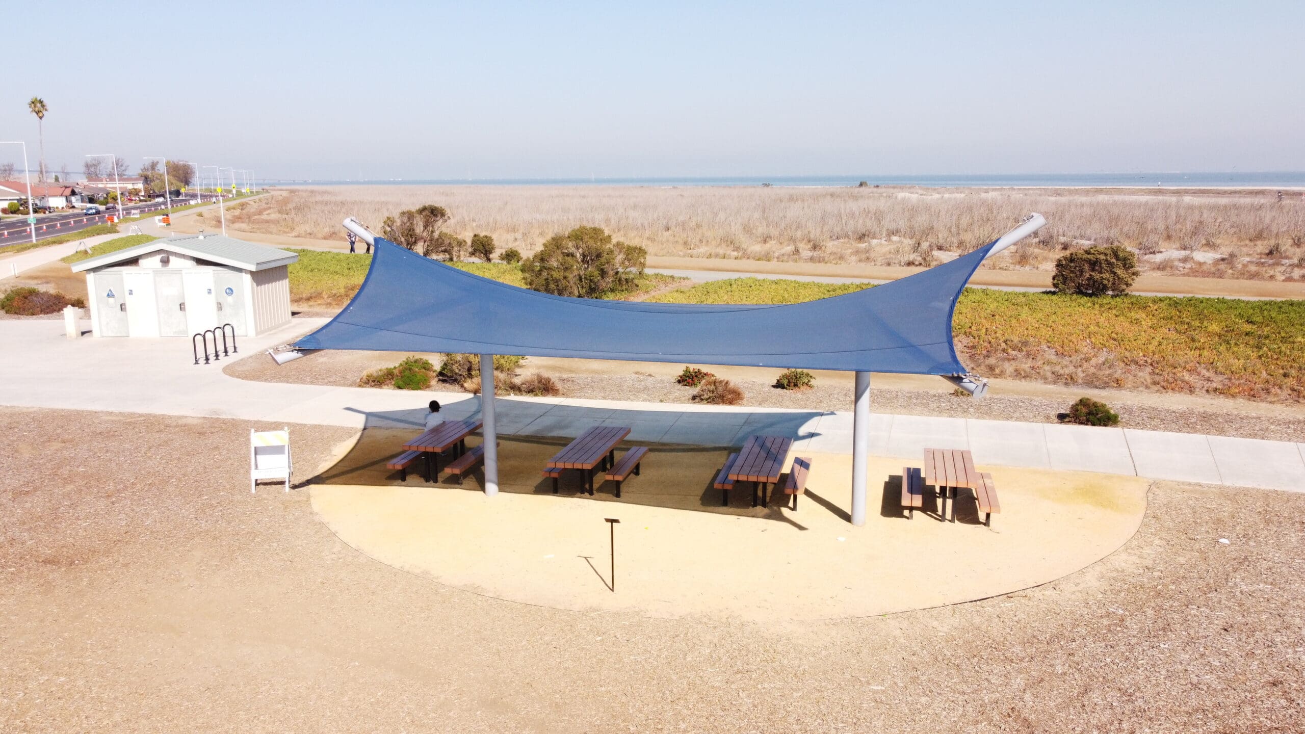A large blue USA Shade Aquarius Shade sail over a Picnic area with picnic tables and chairs with beach in background