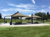 Two large beige USA Shade Mariner Pyramid Shades over picnic tables in a park setting with green grass and trees.