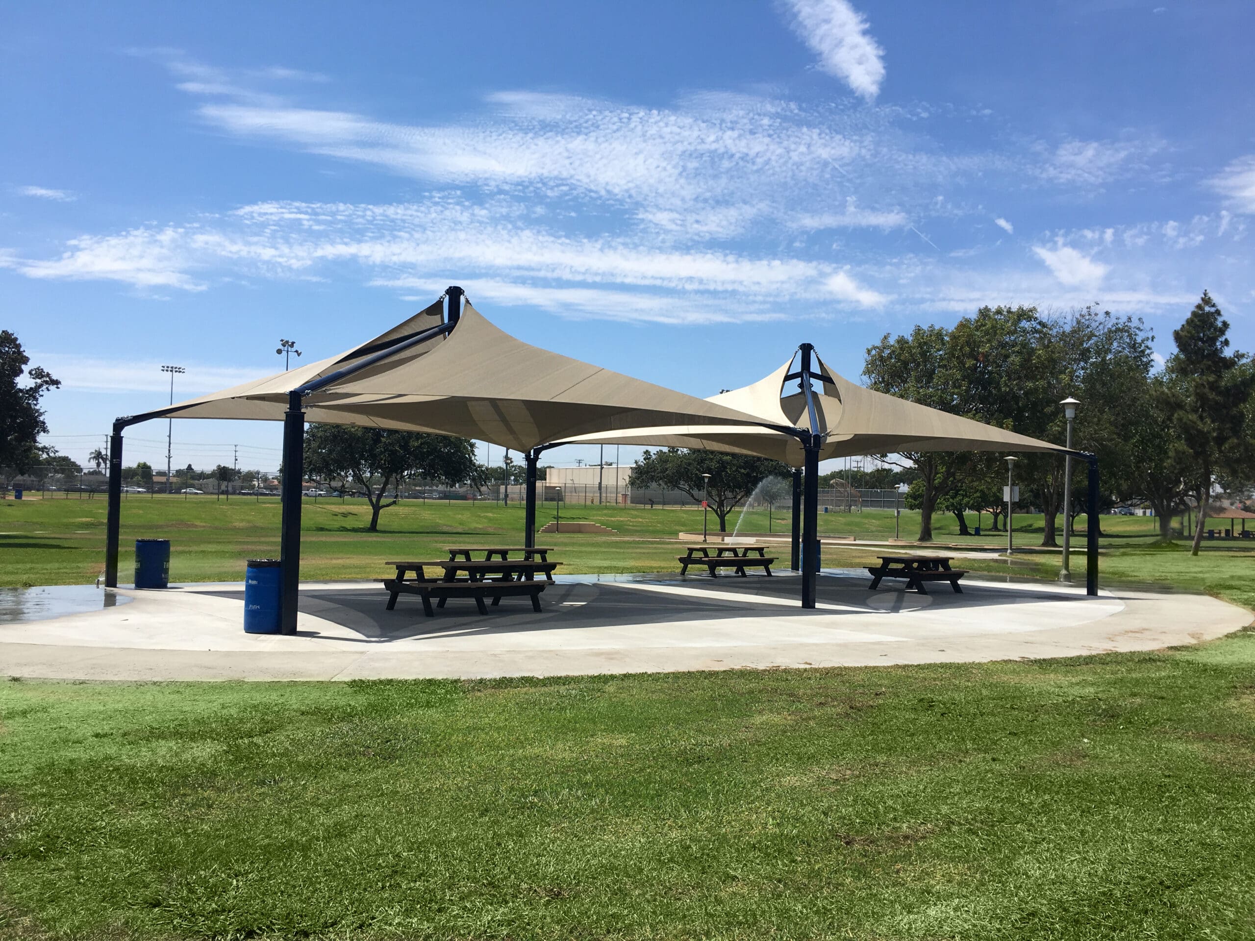 Two large beige USA Shade Mariner Pyramid Shades over picnic tables in a park setting with green grass and trees.