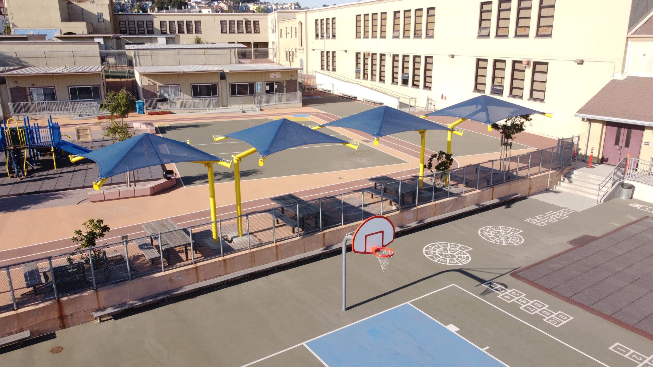 School playground with blue USA Shade Single Post Cantilever Shade Structures, basketball hoop, and outdoor furniture.