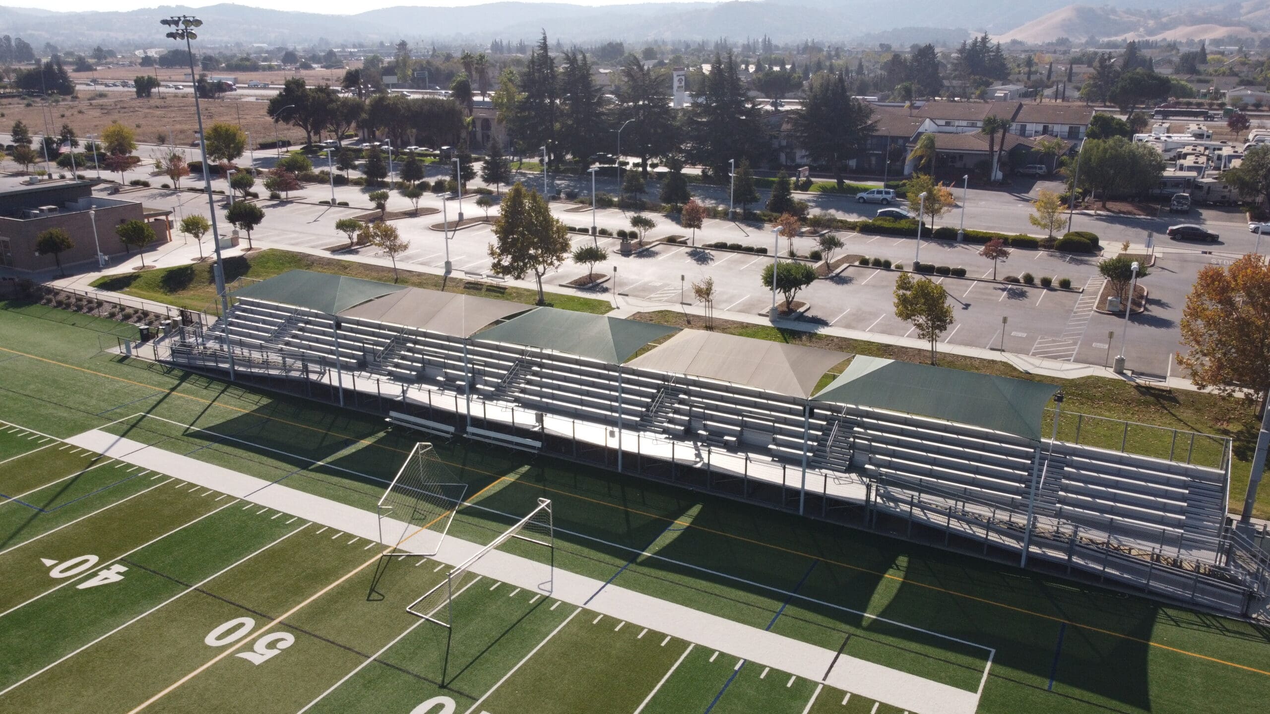 A football field with bleachers covered by USA Shade Hips and a parking lot in the background