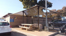 Picnic area with a USA Shade Two Post Hip Shade Structure, tables, and benches under a clear sky.