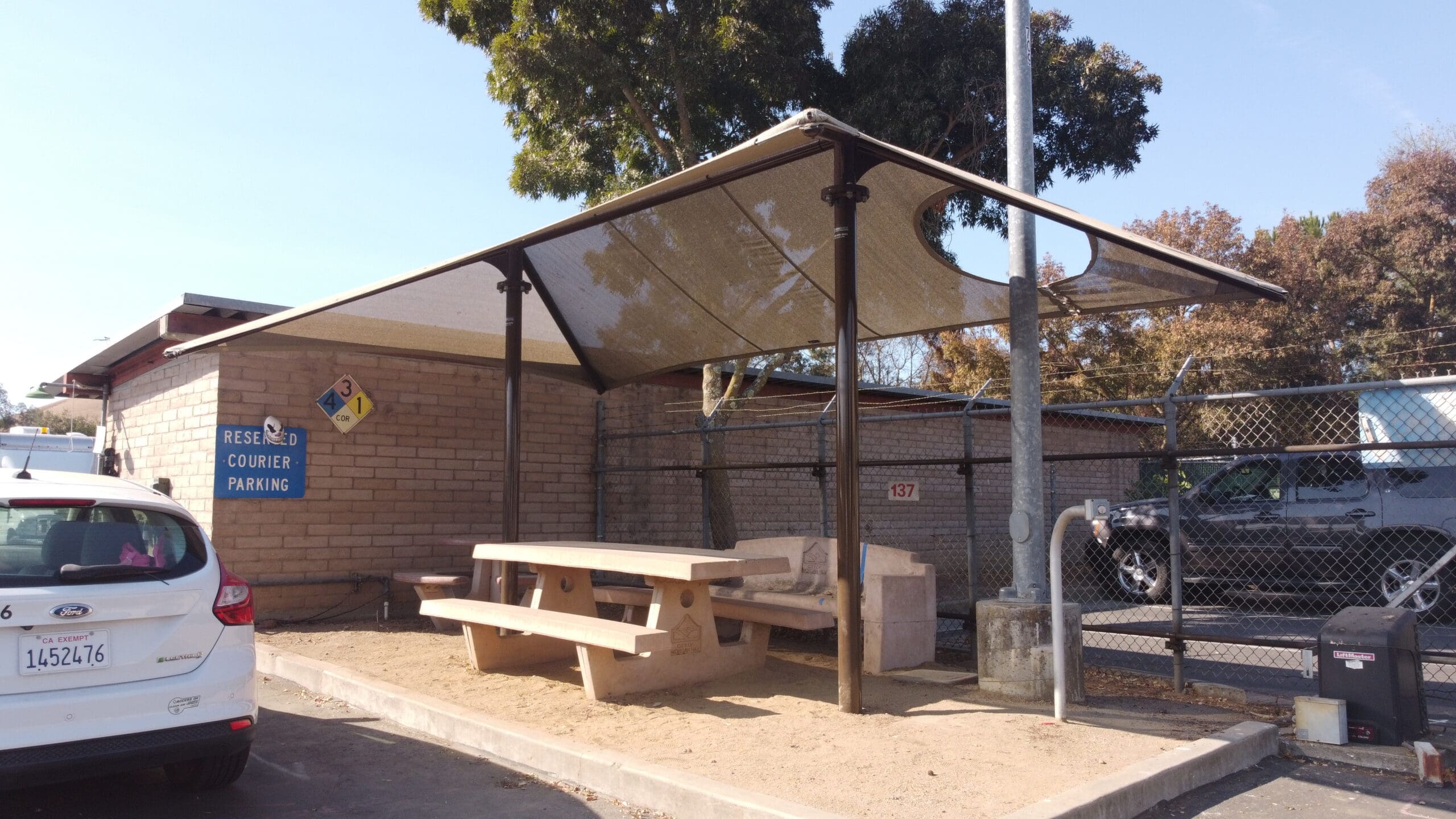 Picnic area with a USA Shade Two Post Hip Shade Structure, tables, and benches under a clear sky.
