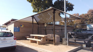 Picnic area with a USA Shade Two Post Hip Shade Structure, tables, and benches under a clear sky.