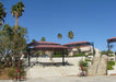 Outdoor seating area with tables covered with USA Shade Tri-Truss Cantilevers and palm trees under a clear blue sky.
