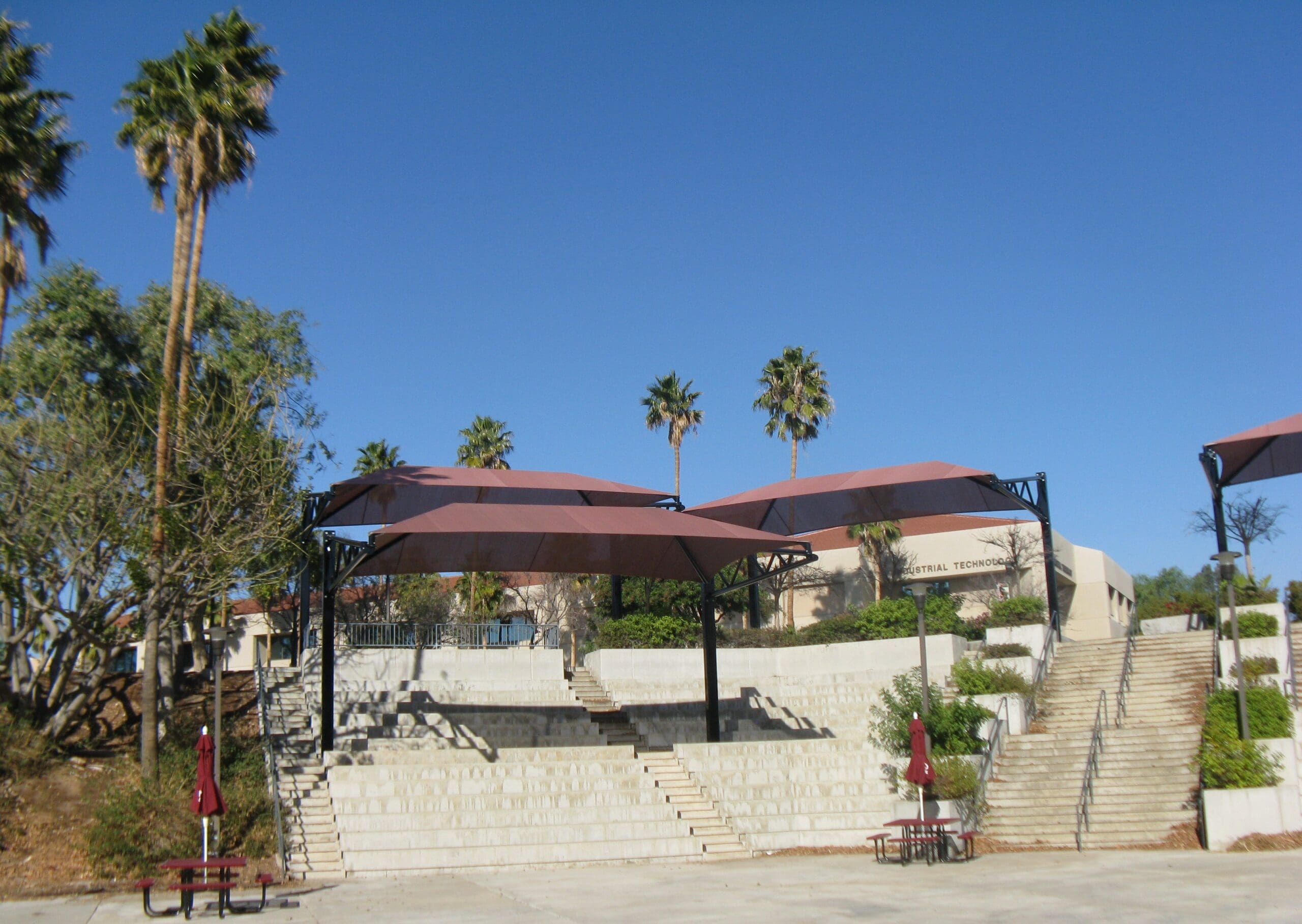 Outdoor seating area with tables covered with USA Shade Tri-Truss Cantilevers and palm trees under a clear blue sky.