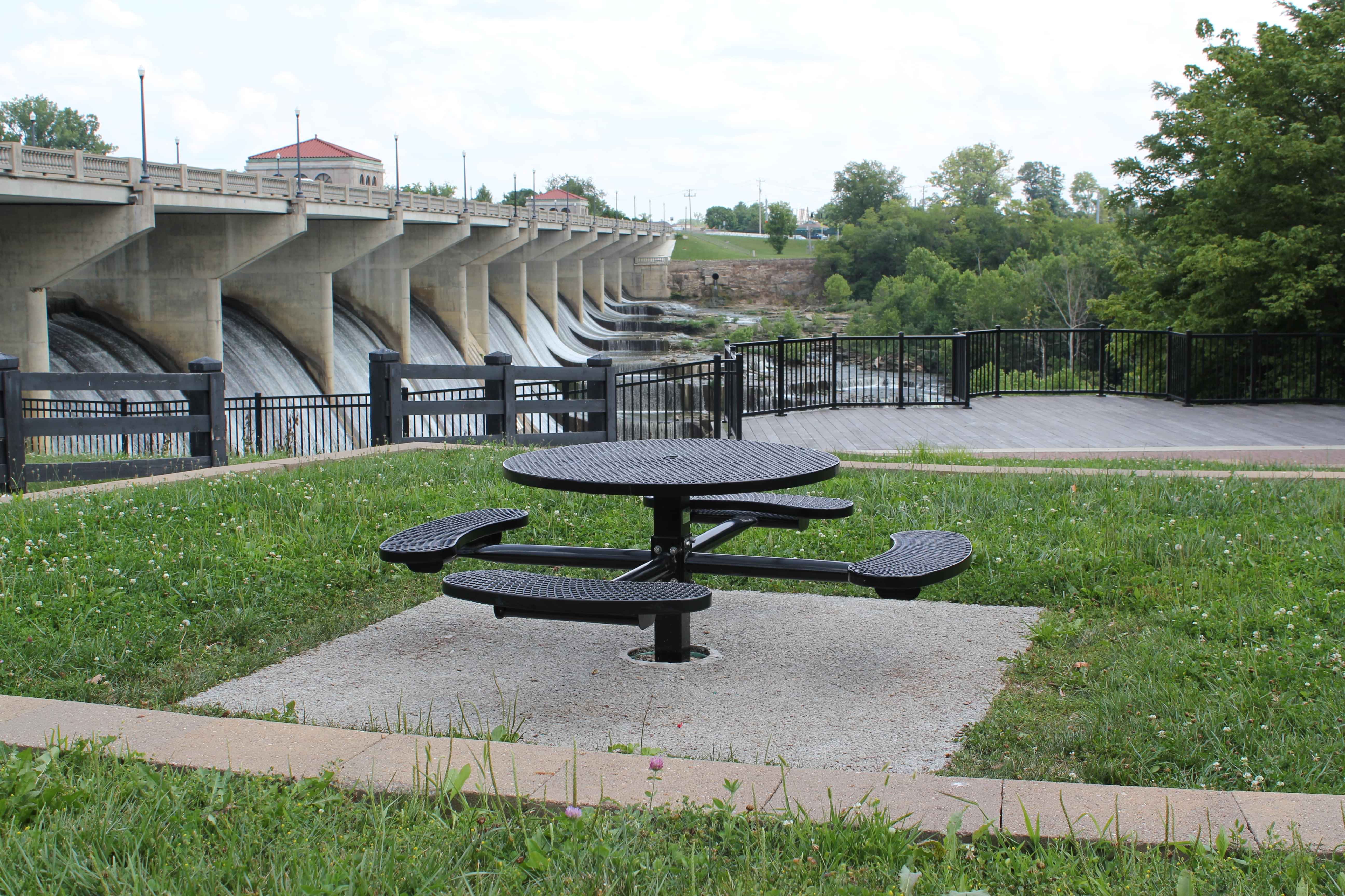 Black Wabash Valley Signature Round Pedestal Picnic Table with a Diamond top and four seats, set inground on a concrete pad in a riverside park, commercial patio furniture