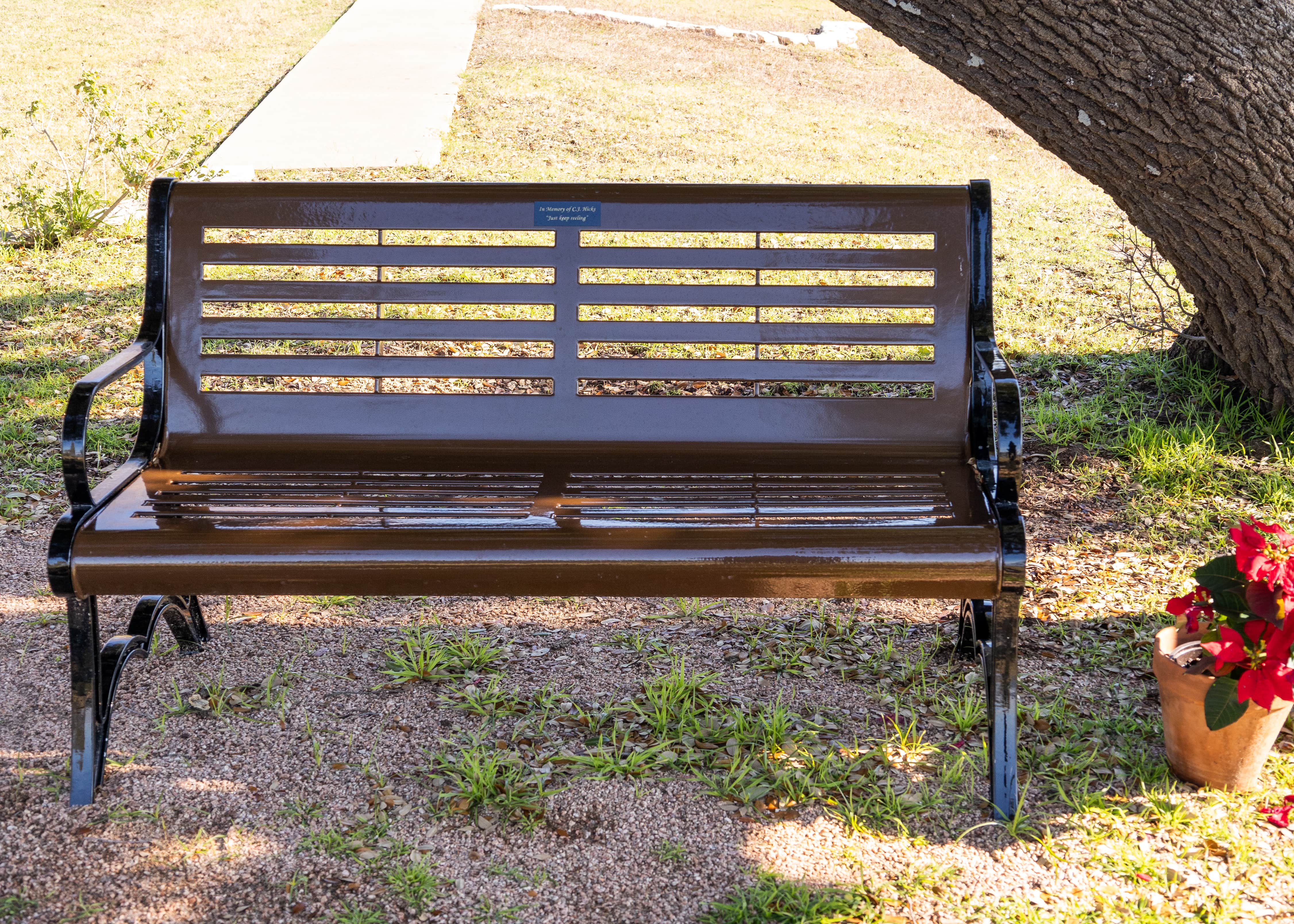Front Angle of Brown MyTCoat Austin Bench with Horizontal Slatted Steel with Park Background and Colorful Flower Pot