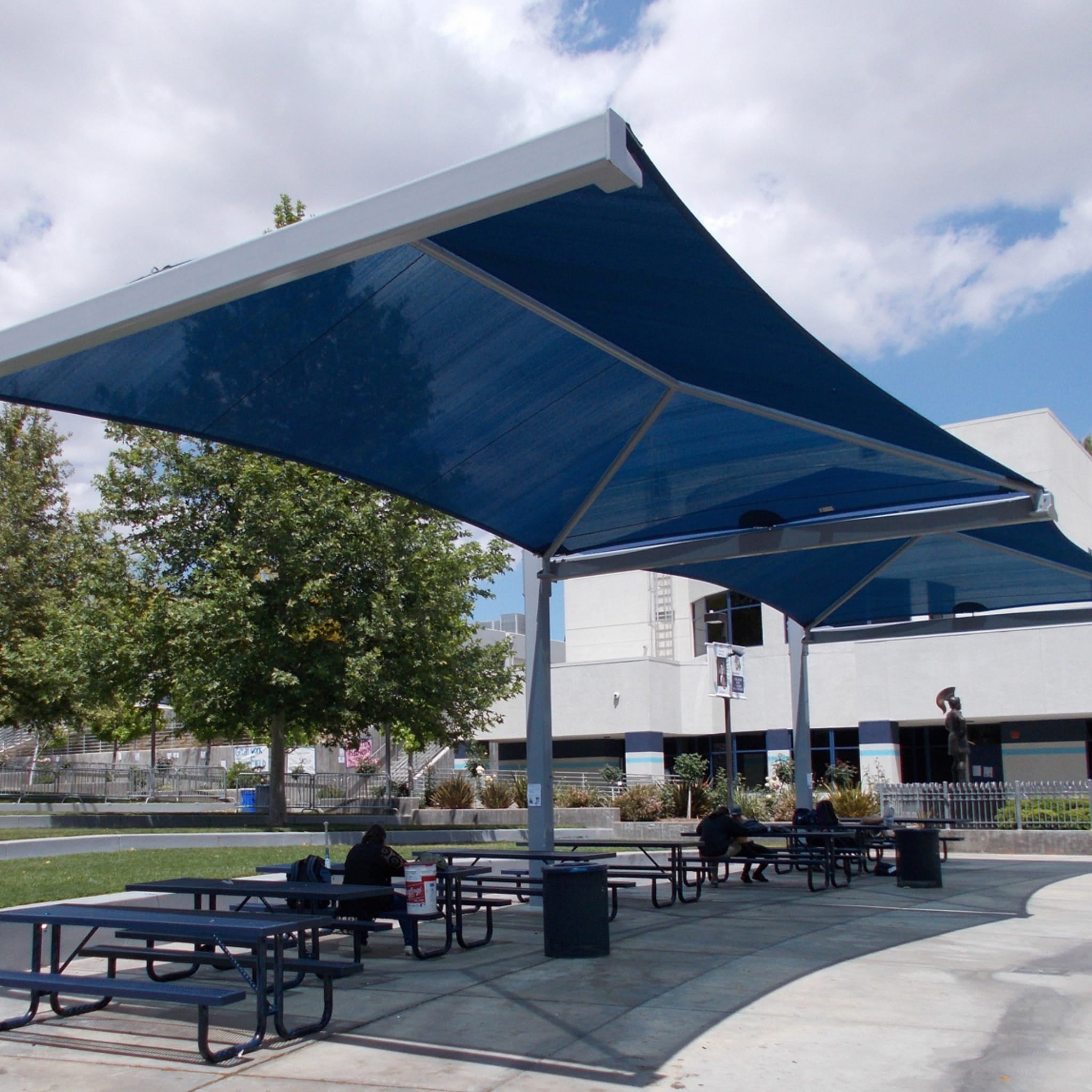 Outdoor seating area with blue USA Shade Full Hip Cantilever Shade, picnic tables, and people sitting under a cloudy sky.