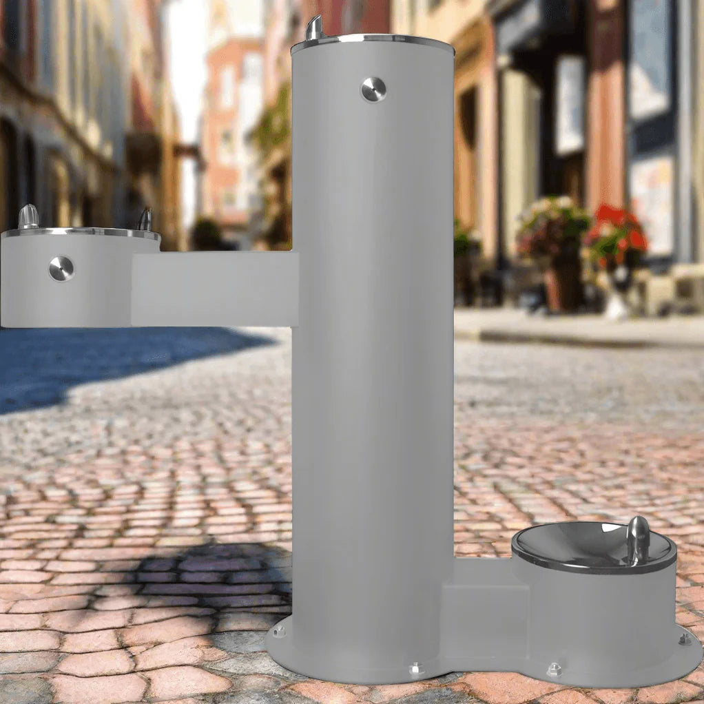 Gray Willoughby Free-standing Double Drinking Fountain Front Access, Child Height, Dog Bowl Options on a cobblestone street with buildings in the background.
