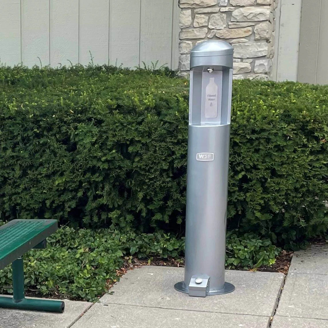Silver Willoughy Outdoor Water Bottle Filling Station with Foot Pedal on a sidewalk next to green bushes