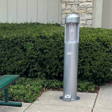Silver Willoughy Outdoor Water Bottle Filling Station with Foot Pedal on a sidewalk next to green bushes