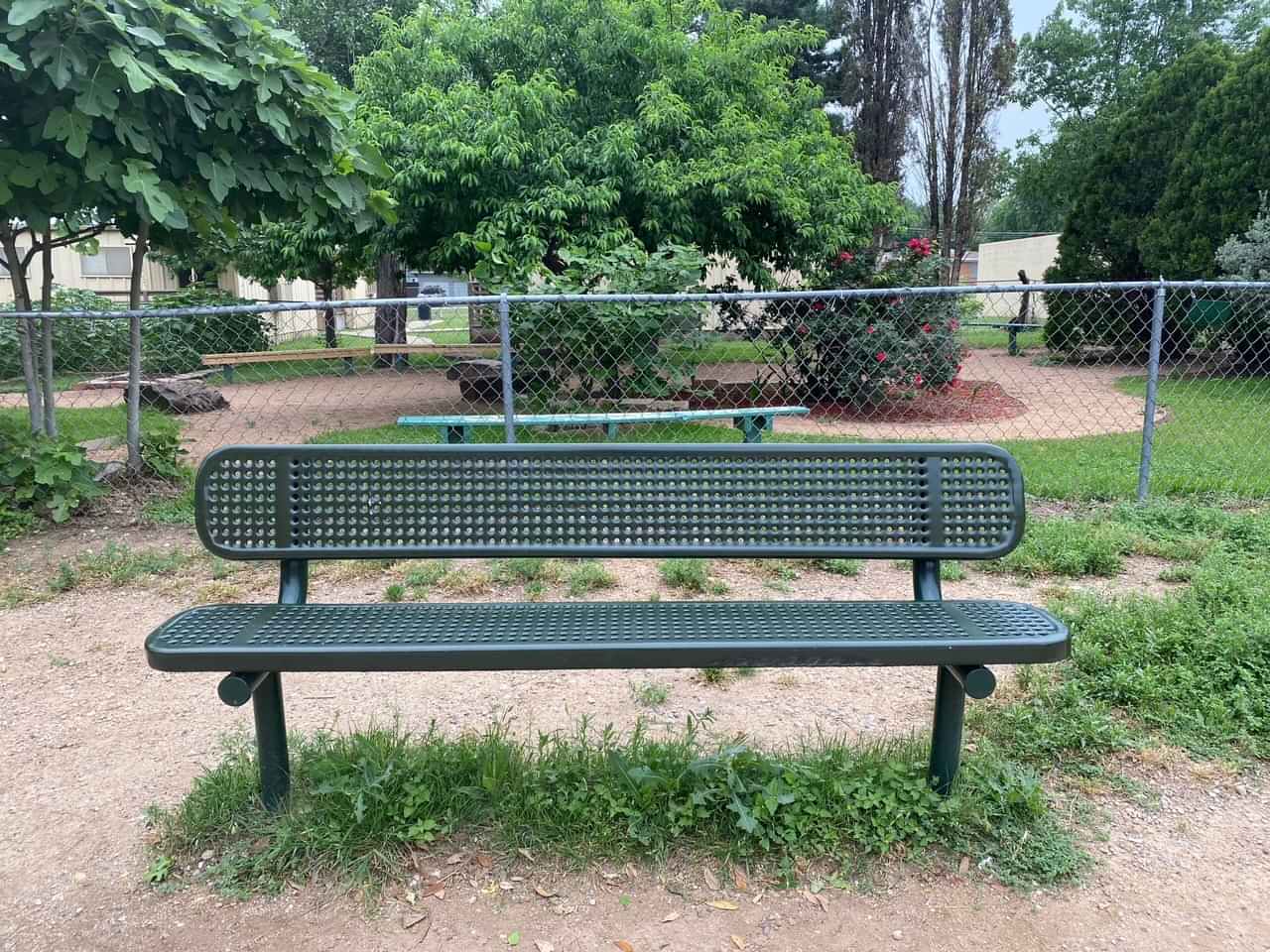 Green Wabash Valley Signature Steel Bench, Perforated seat and back, Inground legs, beside a schoolyard fence, inground bench