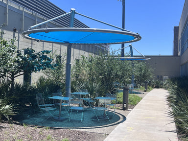 Outdoor seating area with tables and chairs under a large blue USA Shade Cantilever Disc on a sunny day.