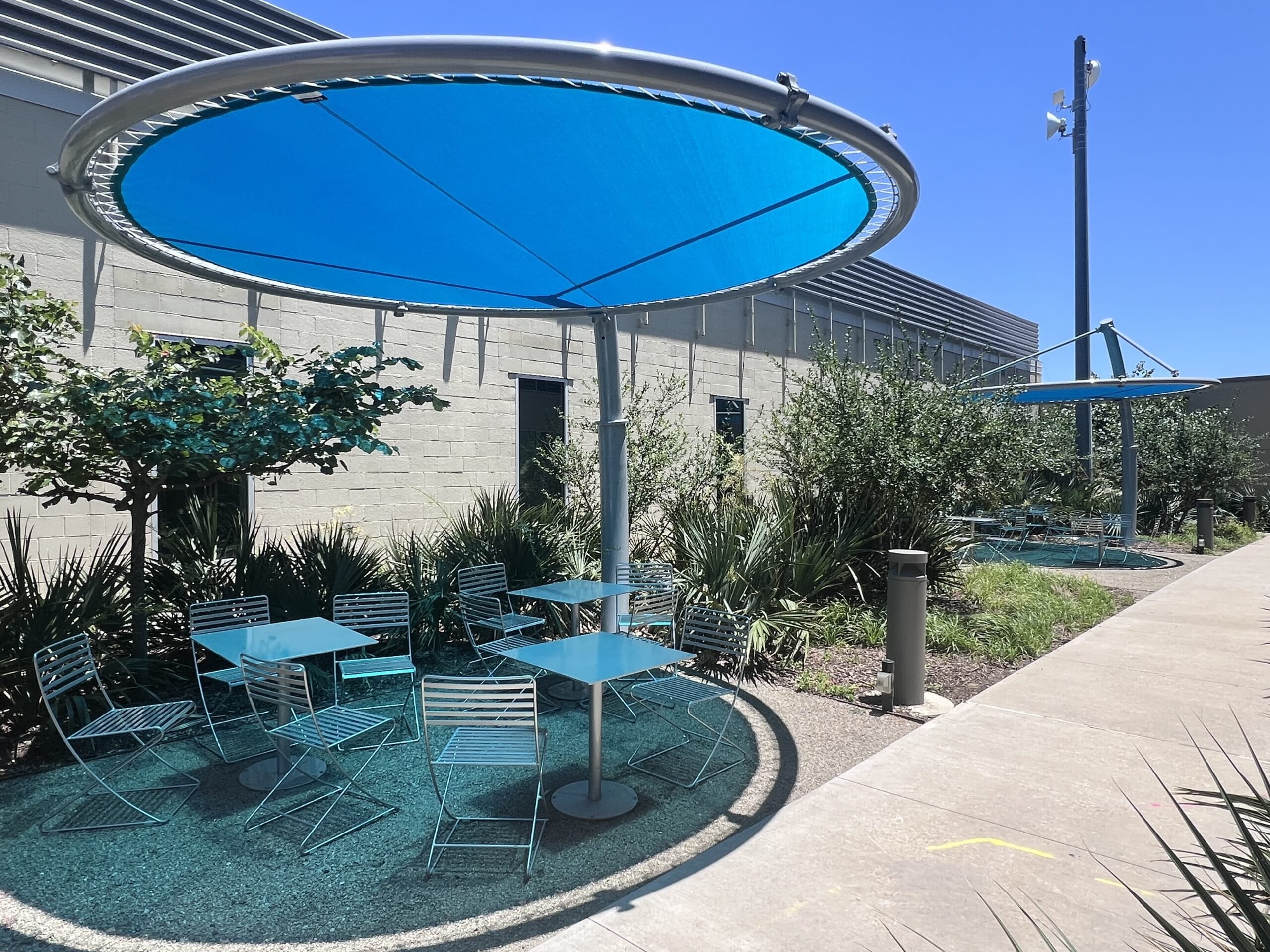 Outdoor seating area with blue USA Shade Cantilever Disc shade structures and tables under a clear blue sky.