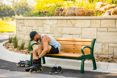 Person sitting on a Frog Furnishings Recycled Plastic Heritage Garden Bench putting on roller skates with a scenic background