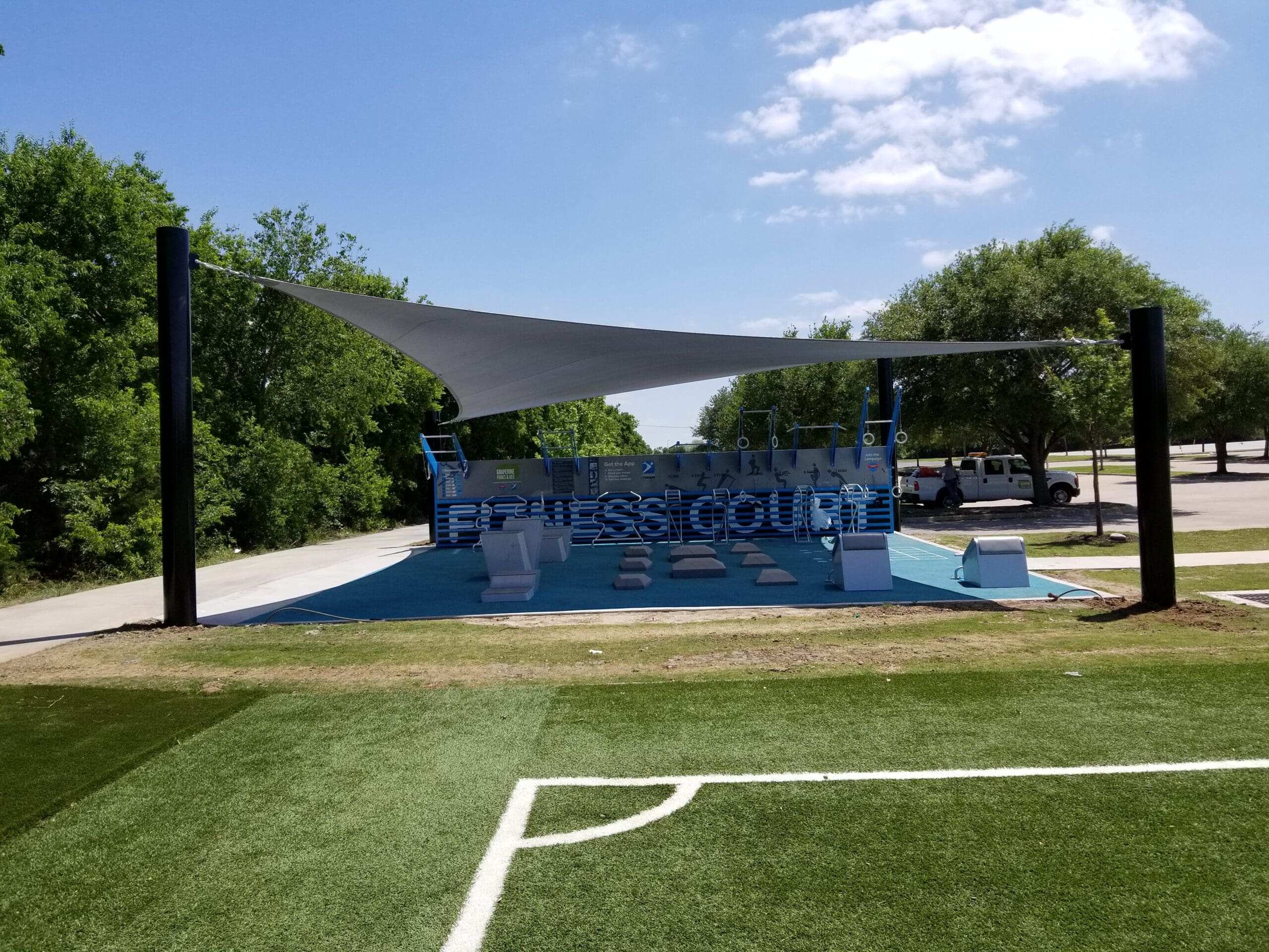 Outdoor sports facility with blue and white equipment under a shade sail, surrounded by trees and a clear sky.