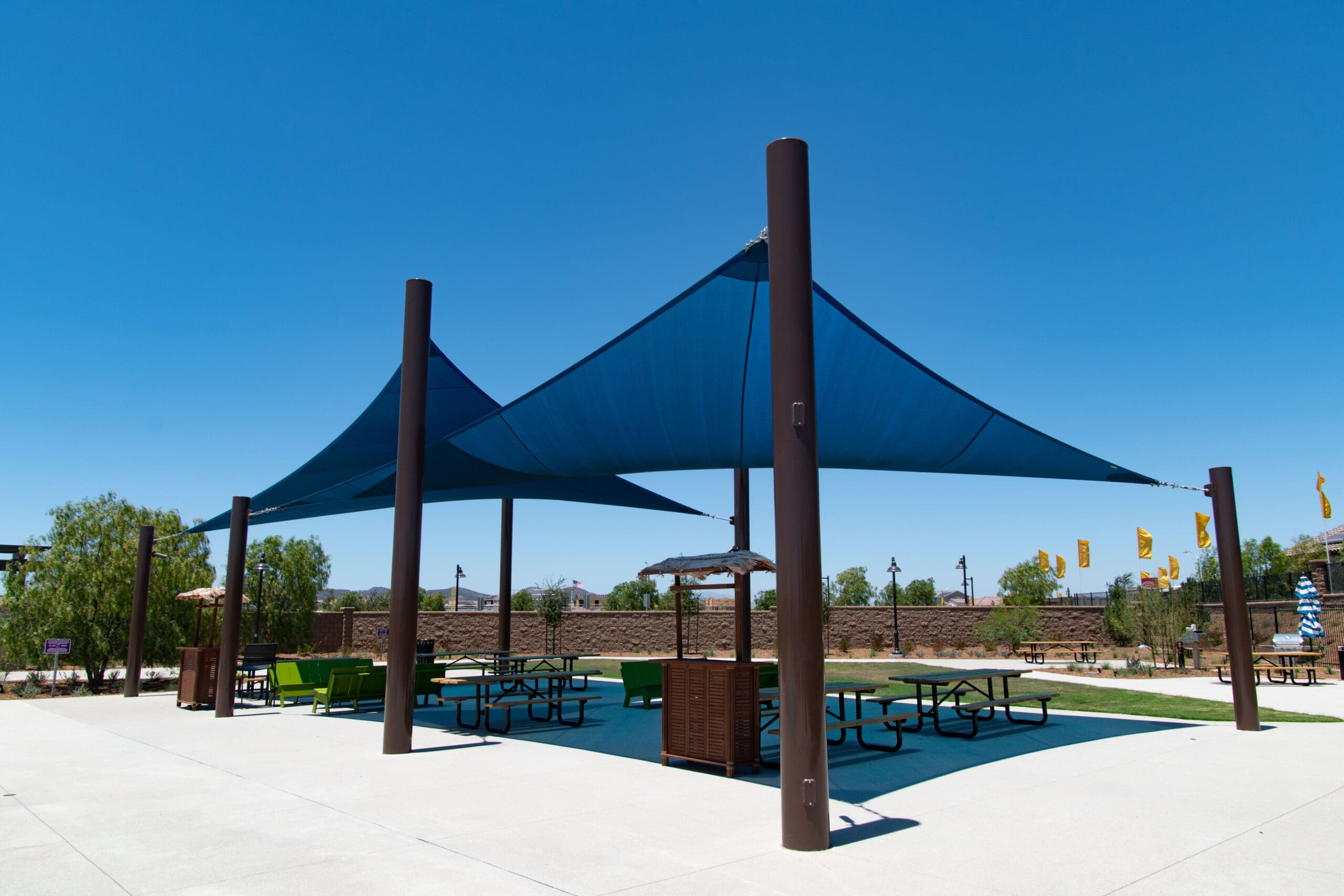 Outdoor shaded area with blue 4-point Hypar sail shades and picnic tables under a clear blue sky.