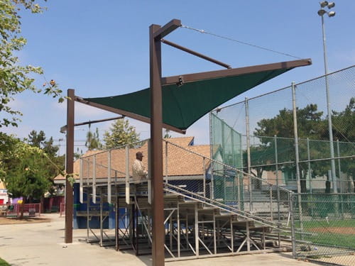 Outdoor setting with a green Cantilever Hypar Sail shade sail over a stage area, surrounded by trees and a fence.