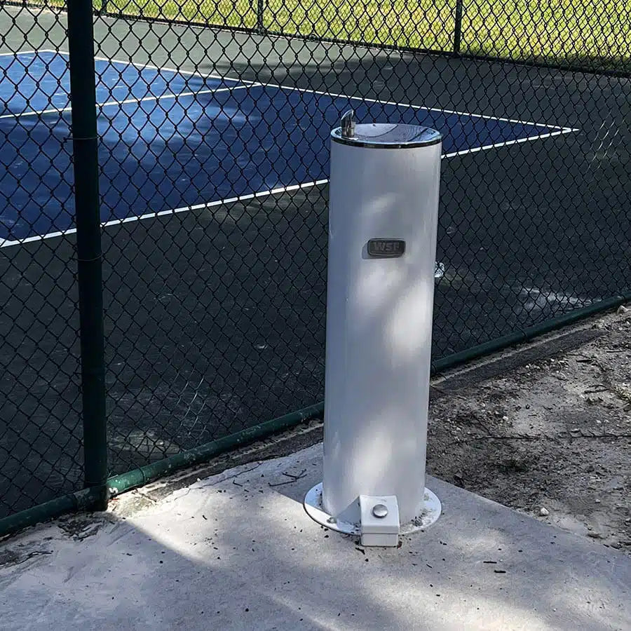 White Willhoughby Single Outdoor Drinking Fountain with Foot Pedal on a concrete pad next to a chain-link fence with a tennis court in the background.