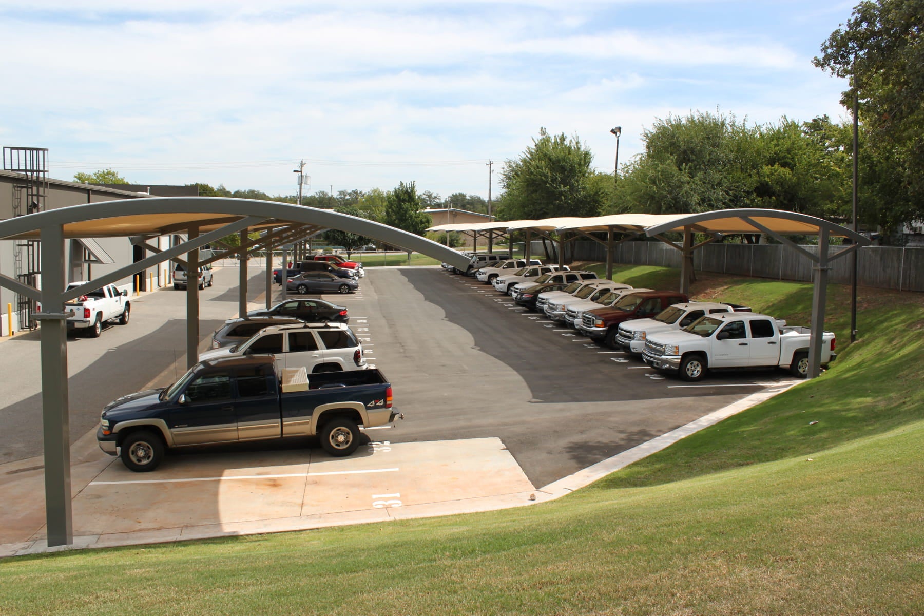 Parking lot with cars and trucks under USA Shade Panorama Cantilever covered areas, surrounded by greenery.