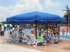 People enjoying a poolside area with a large blue USA Shade Coolbrella, lounge chairs, and a pool in the background.