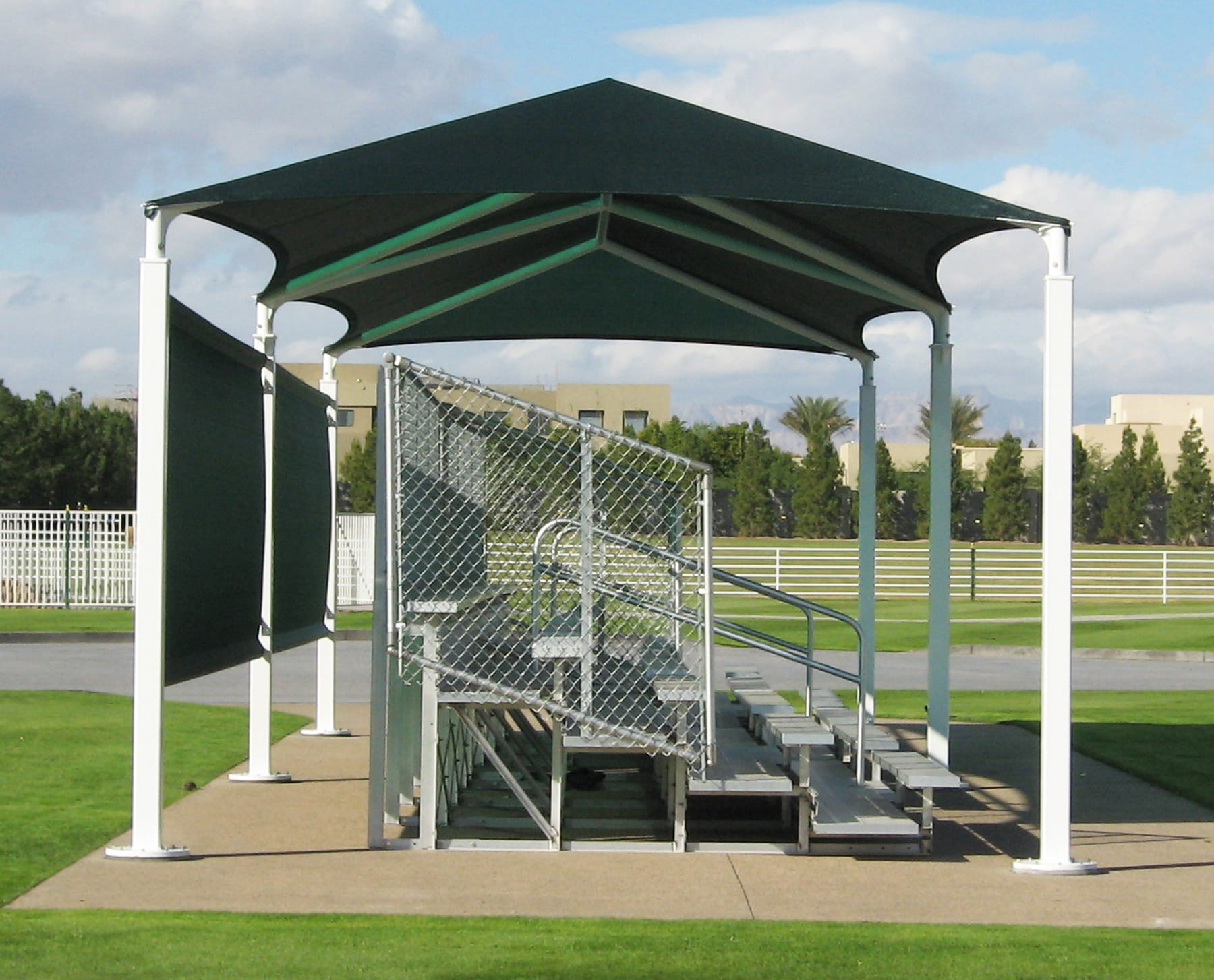 Outdoor sports facility with a green USA Shade 6-Post Hip and metal bleacher leading to a dugout.