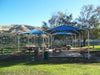 Outdoor picnic area with blue USA Shade Multi-Panel Pyramids by a lake on a sunny day