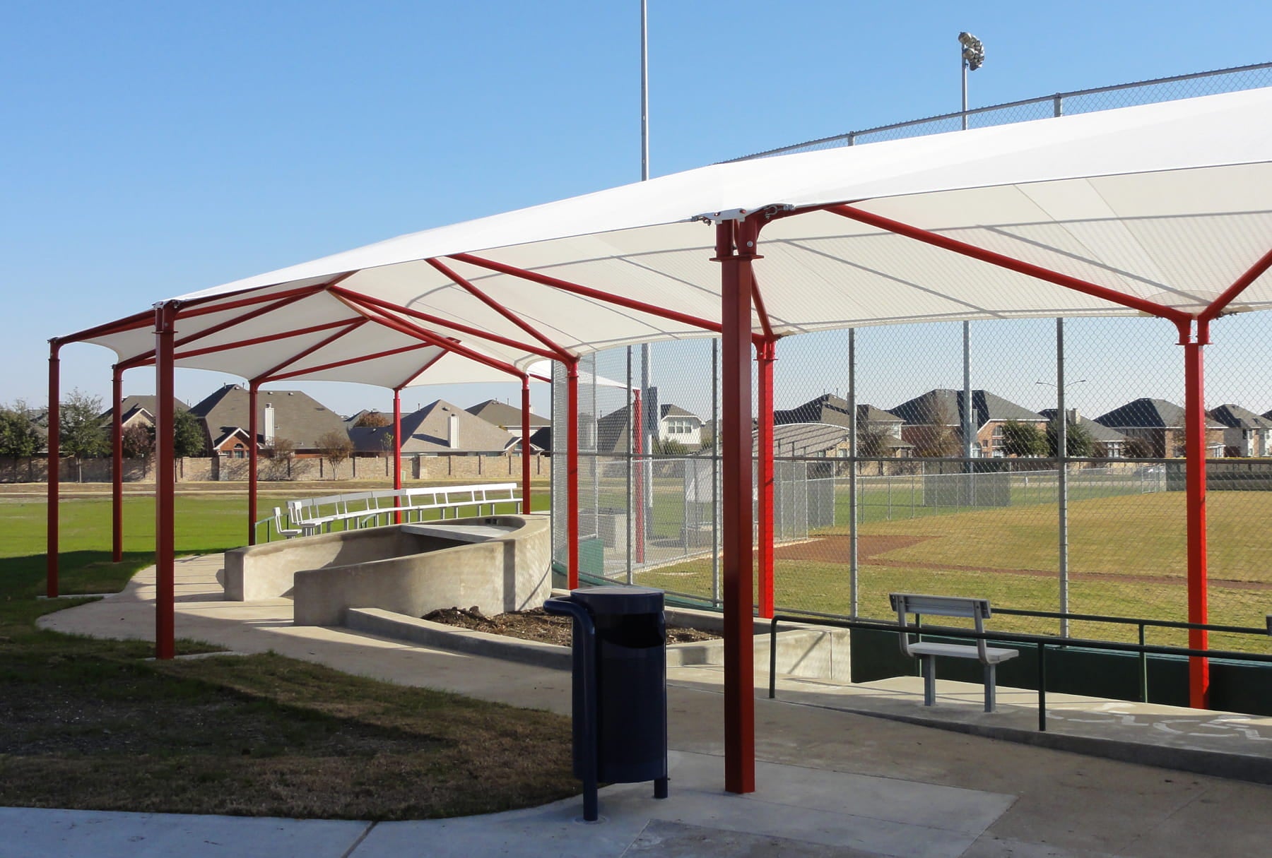 USA Shade Wrap-around Hip covering seating area at a sports field with red frames and white roof, surrounded by a fence and grass.