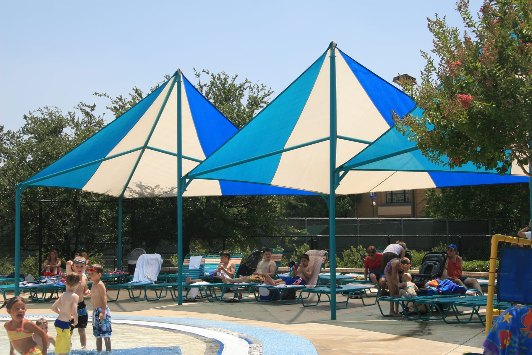 People relaxing under blue and white USA Shade Wave Shades by a poolside.