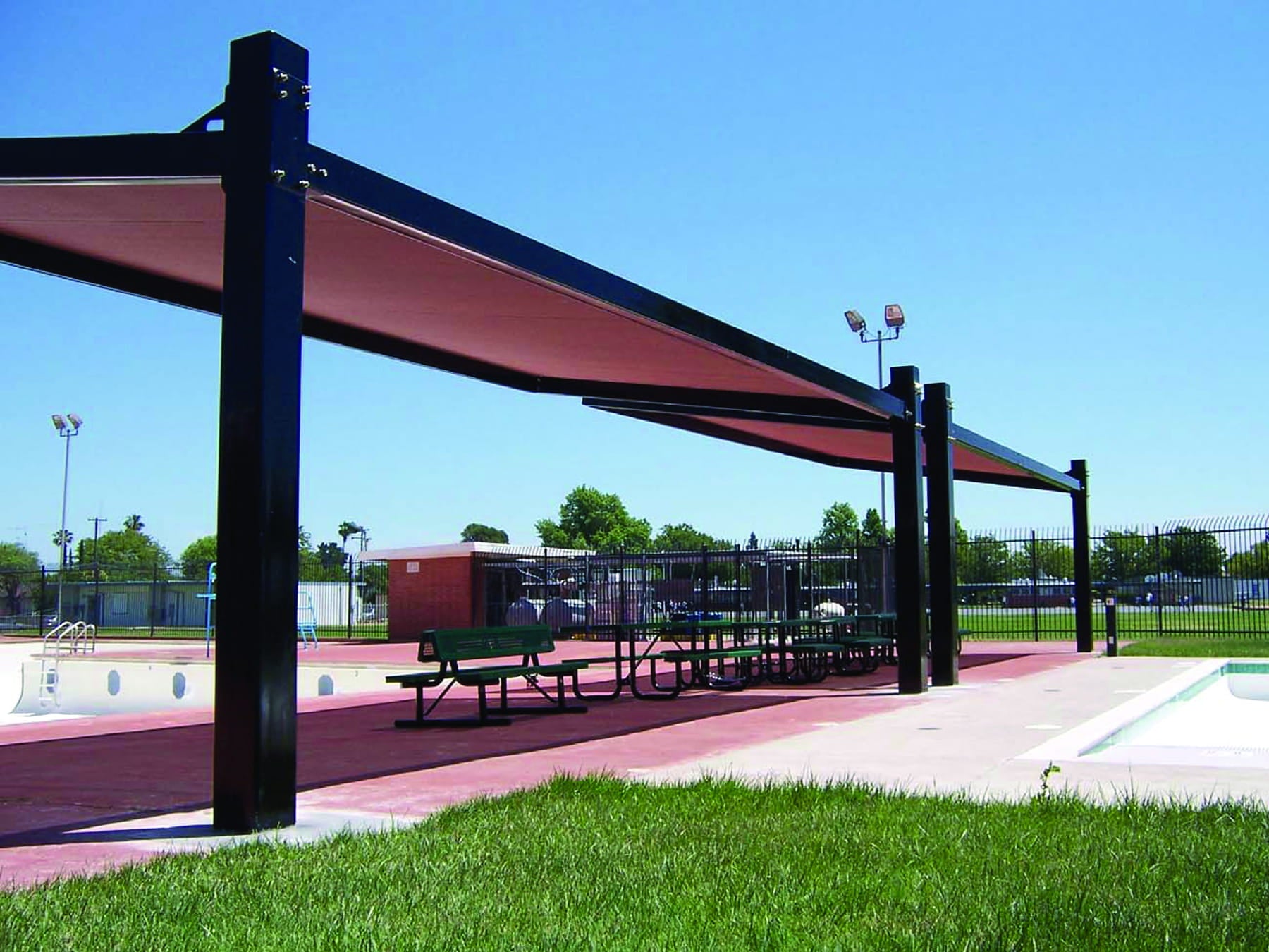 Outdoor sports facility with a USA Shade Slanted Cantilever Framed covered area, benches, and sports equipment under a clear blue sky.