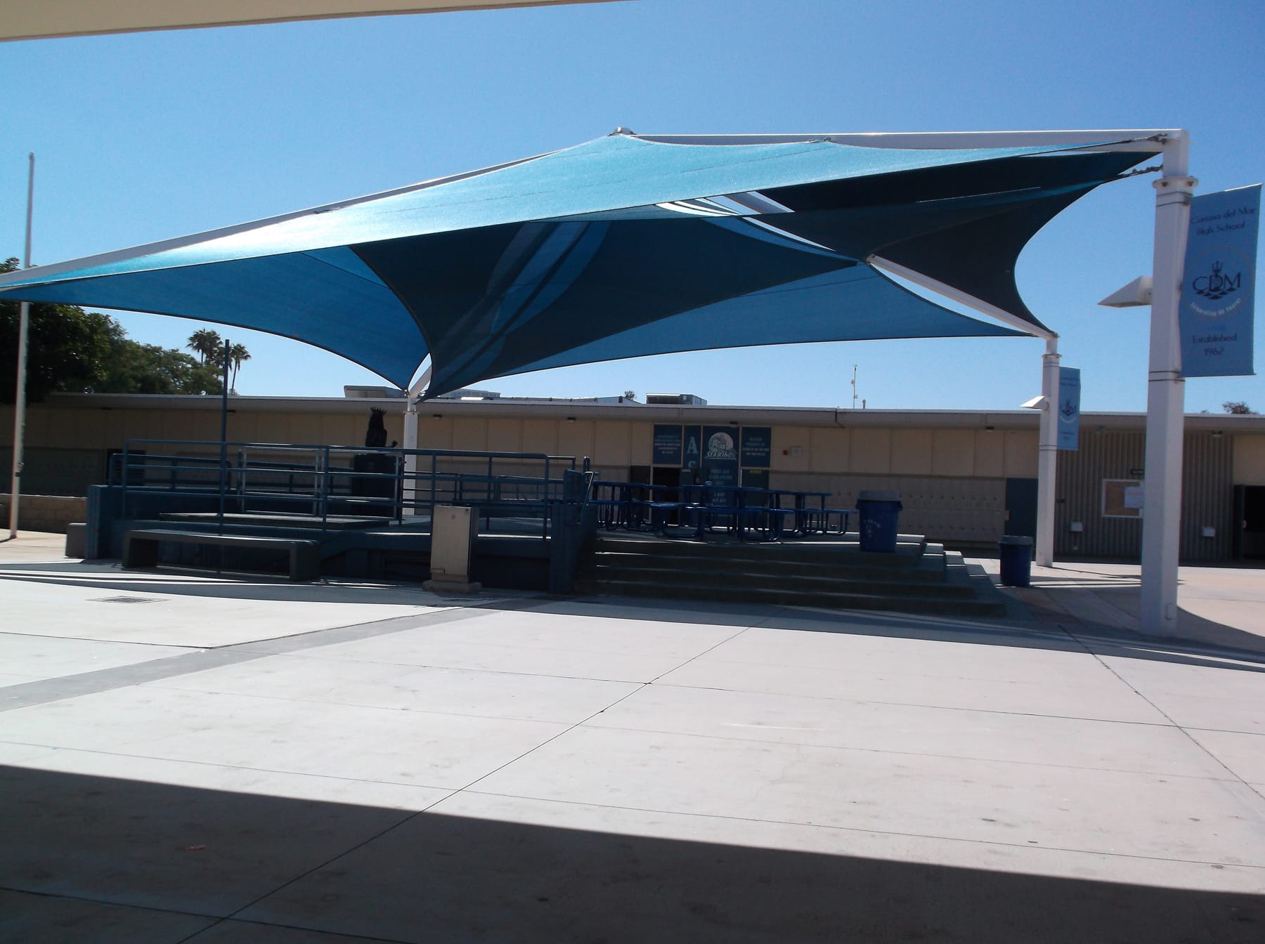 Large blue USA Shade Mariner Pyramid Shade over a concrete area with a school building in the background