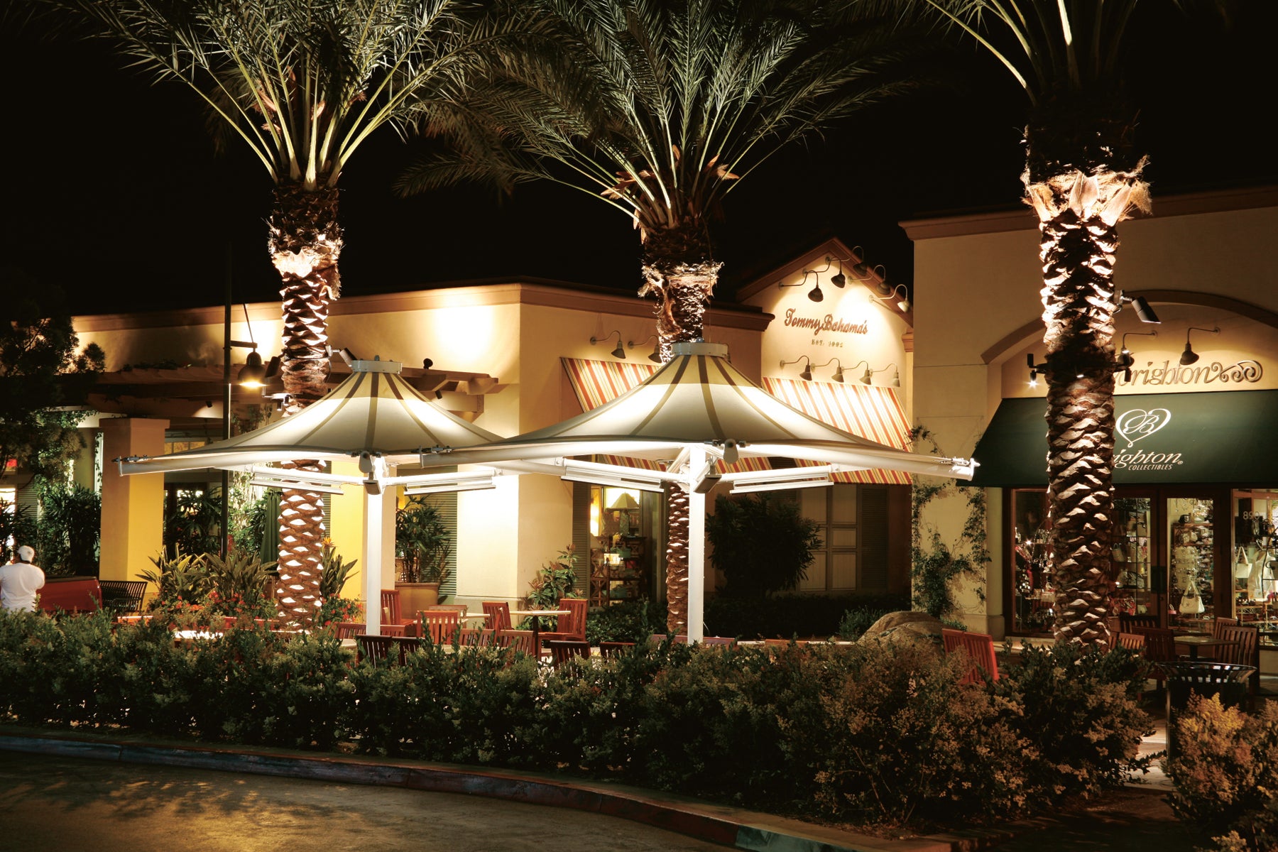 Evening view of a restaurant exterior with USA Shade 4-point Twilite Umbrellas, palm trees and illuminated sign.