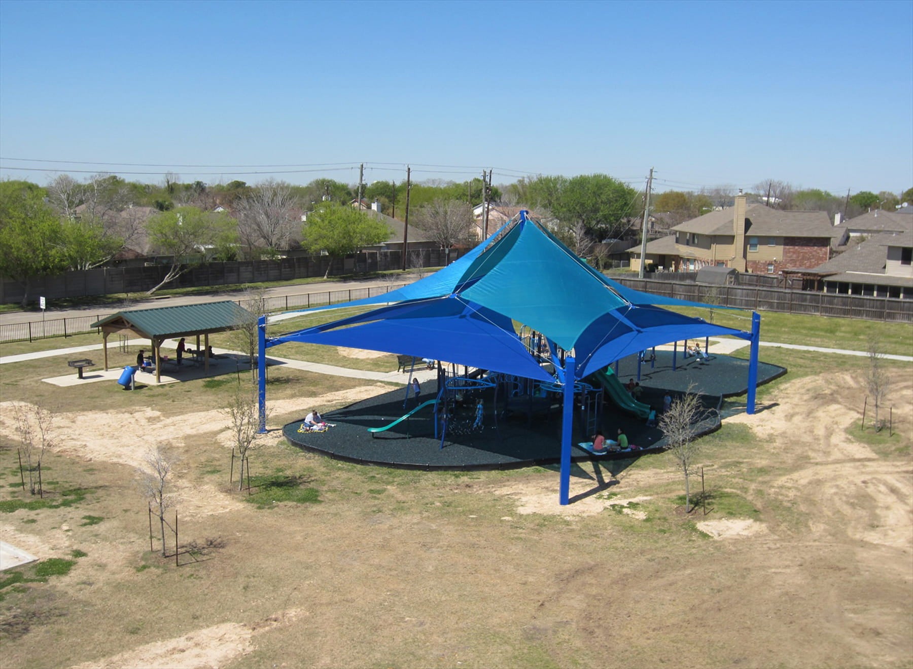 Blue USA Shade Multi-layer Super Span Pyramid Shade structure in a park with trees and buildings in the background